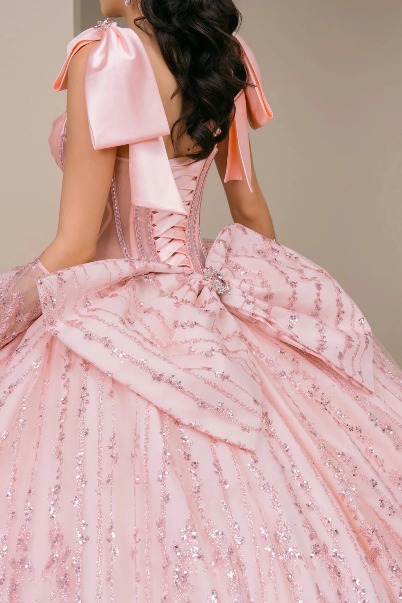 Close-up of woman back-facing in blush off-shoulder sweetheart quinceañera dress with lace-up beaded corset bodice, lace appliqué, oversized back bow, and layered tulle skirt detail