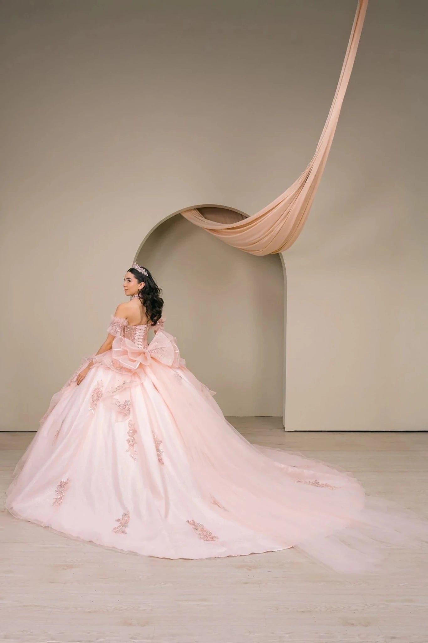 Woman wearing blush off-shoulder sweetheart quinceañera dress three-quarter view with ornate beaded corset bodice, floral appliqué, oversized bow, and voluminous train silhouette