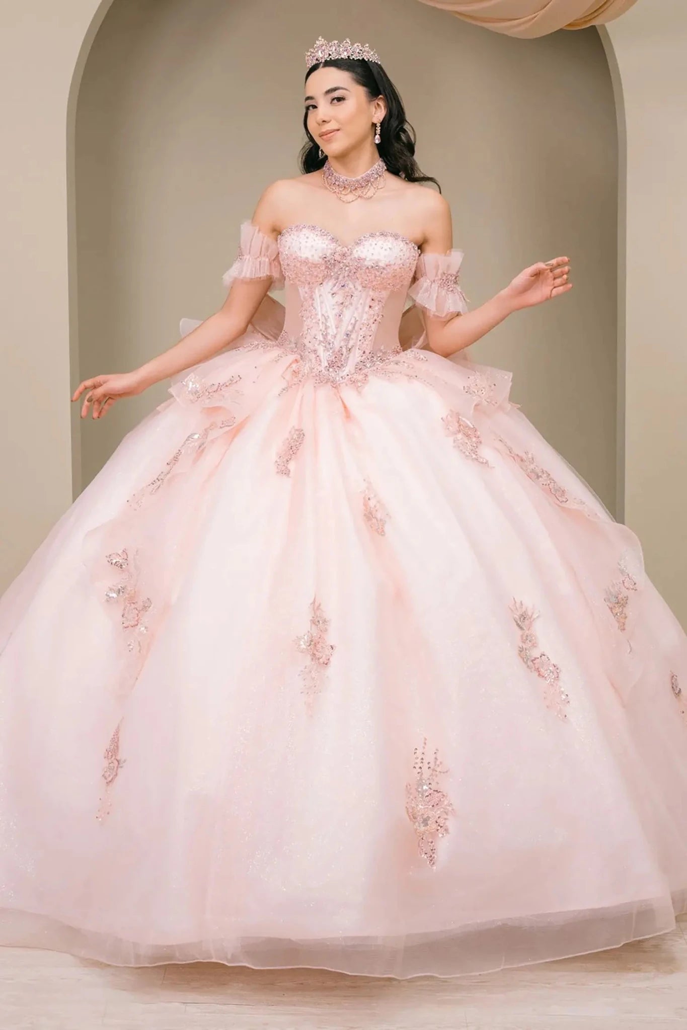 Woman wearing blush off-shoulder sweetheart quinceañera dress with beaded corset bodice, floral appliqué, oversized bow, and flowing train