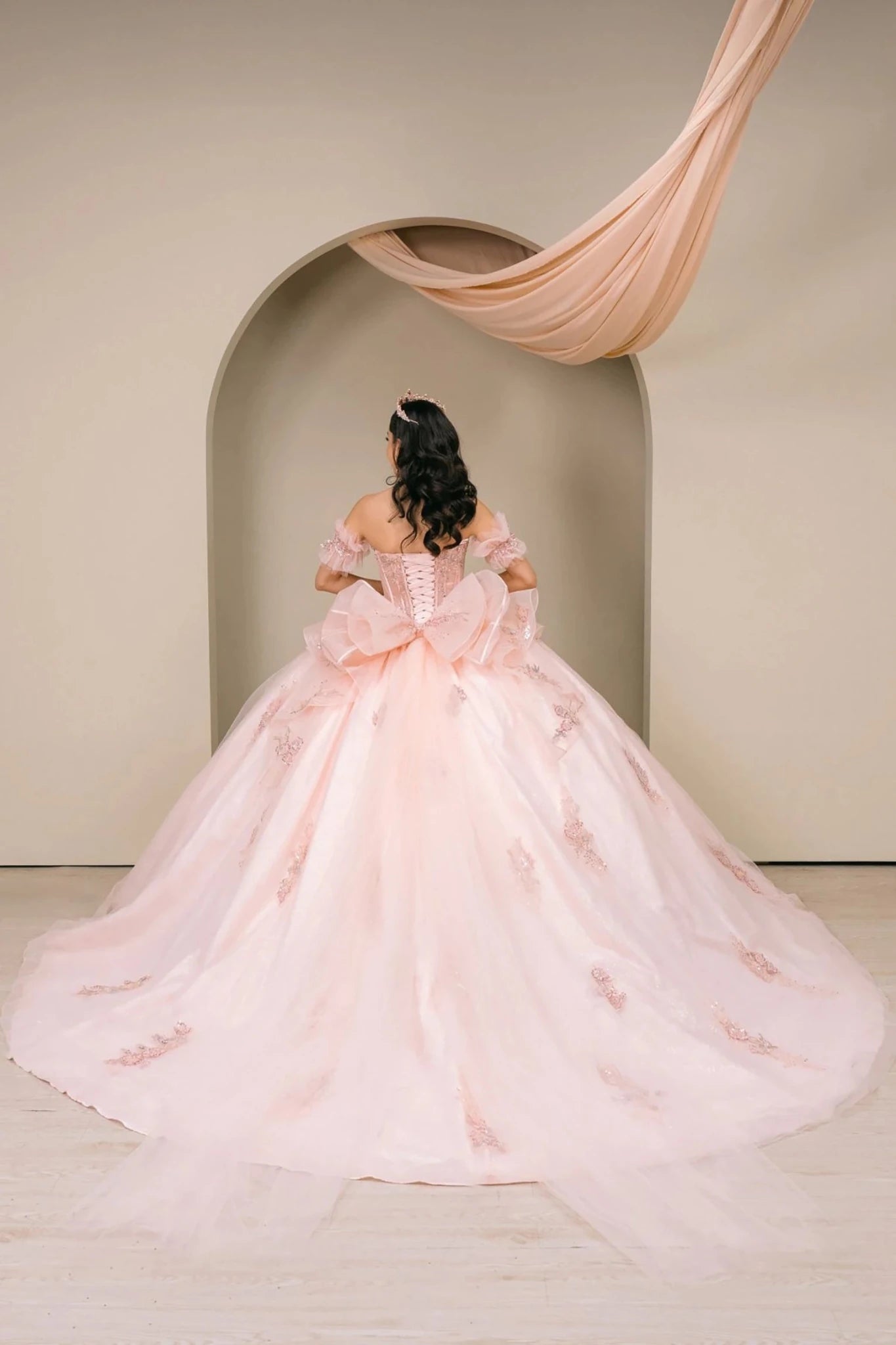 Woman back-facing in blush off-shoulder sweetheart quinceañera dress with lace-up beaded corset bodice, oversized bow detail, floral appliqué, and dramatic train