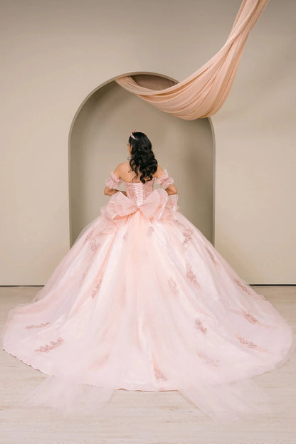 Woman back-facing in blush off-shoulder sweetheart quinceañera dress with lace-up beaded corset bodice, oversized bow detail, floral appliqué, and dramatic train