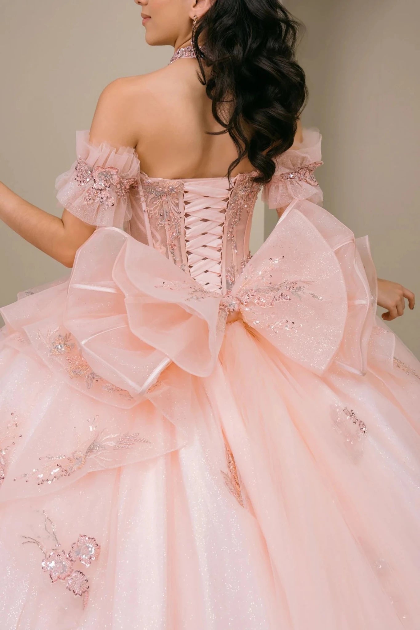 Close-up of woman back-facing in blush off-shoulder sweetheart quinceañera dress with lace-up beaded corset bodice, oversized bow, floral appliqué, and train detail