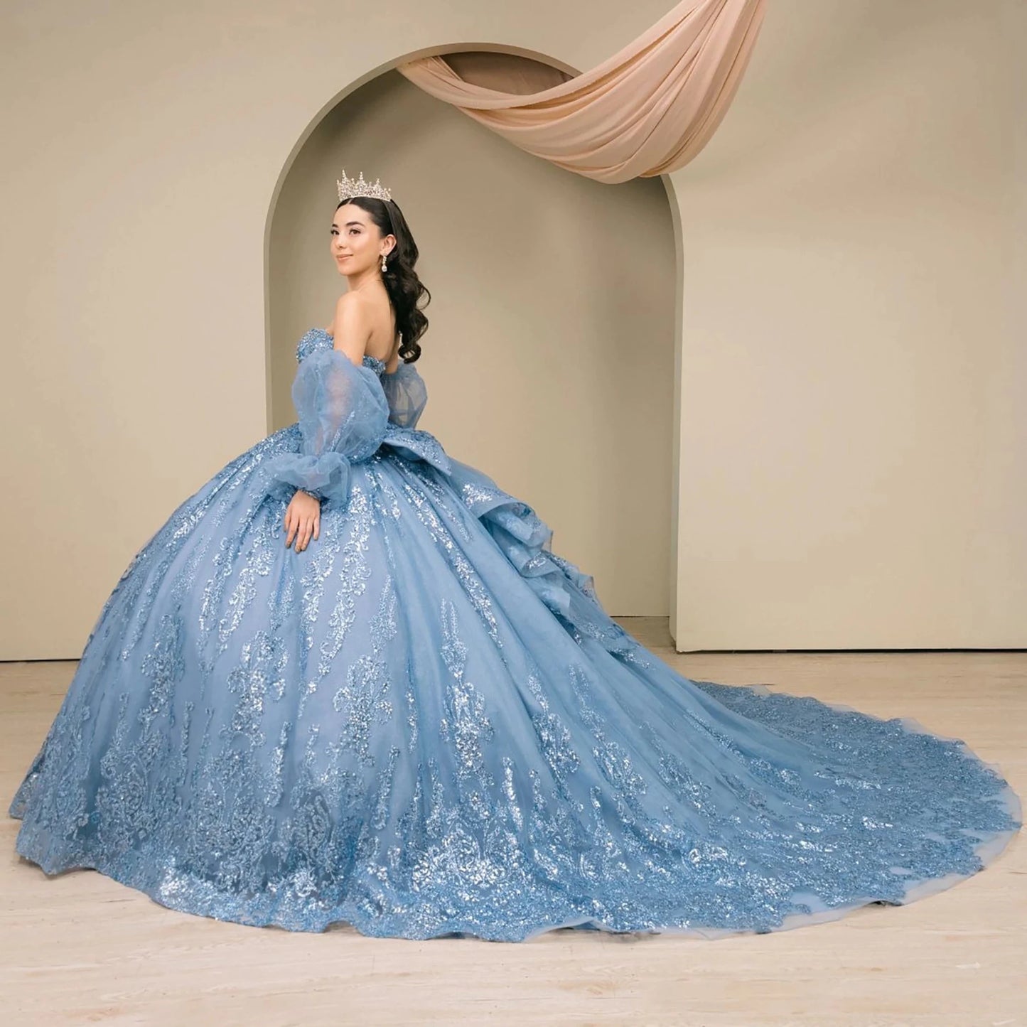 Woman wearing dusty blue off-shoulder sweetheart quinceañera dress side-facing with beaded sequin bodice, voluminous tulle skirt, and flowing sleeve accents