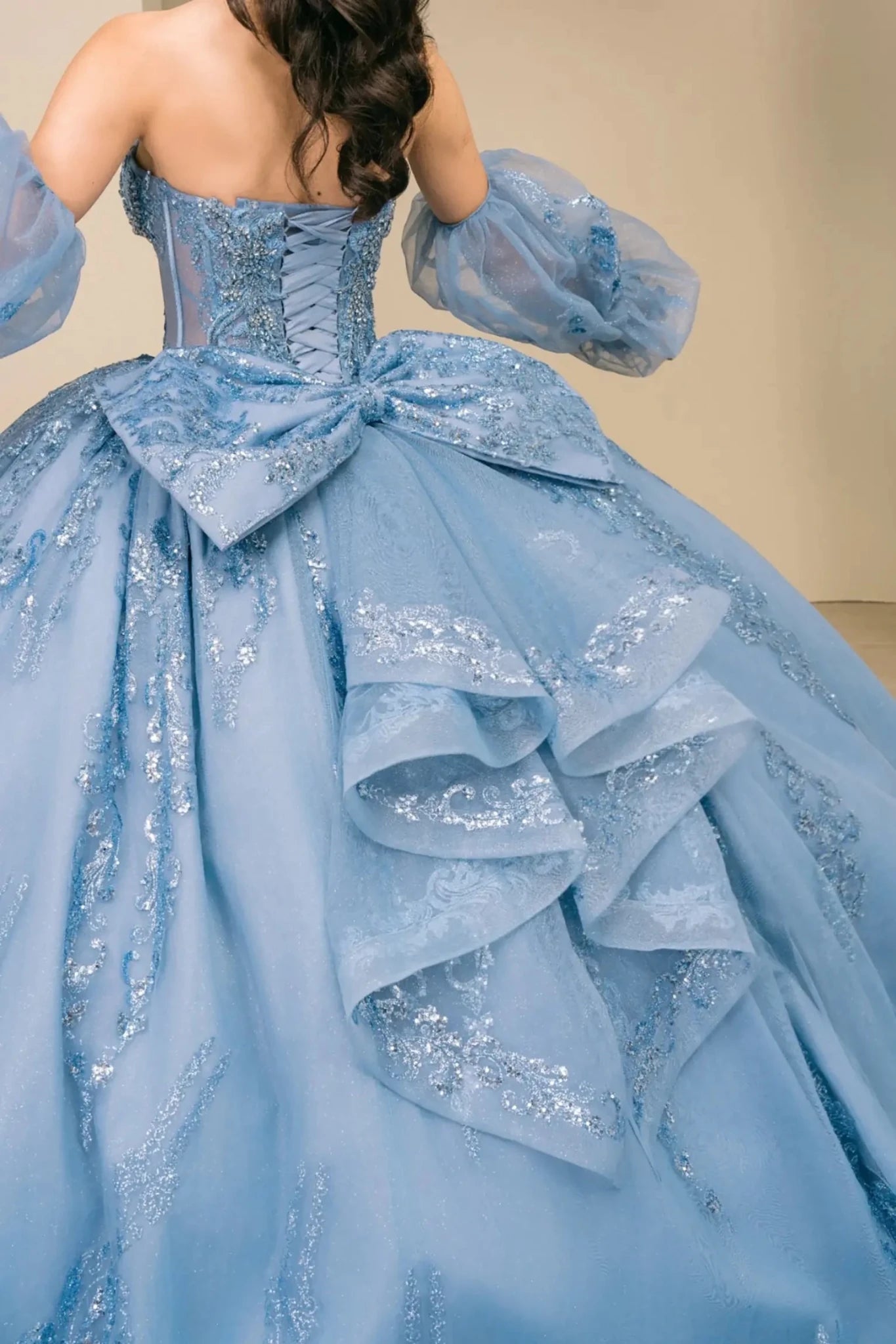Close-up of woman back-facing in dusty blue off-shoulder sweetheart quinceañera dress with lace-up sequin bodice, voluminous tulle skirt, and sleeve embellishments