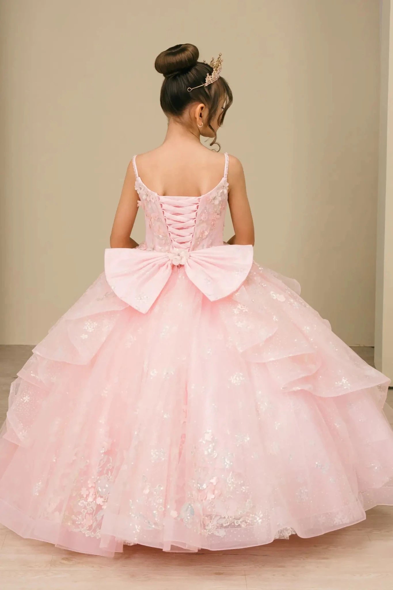 Young girl standing back-facing in mini quinceañera ball gown with lace-up corset bodice, oversized bow accent, and layered skirt