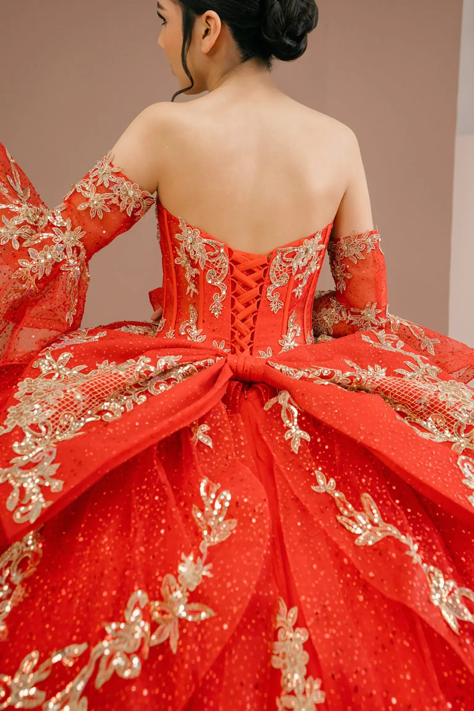 Bodice back close-up of a woman wearing red quinceañera dress with gold embroidery, sequins, and lace-up corset design framed by flowing sleeves.