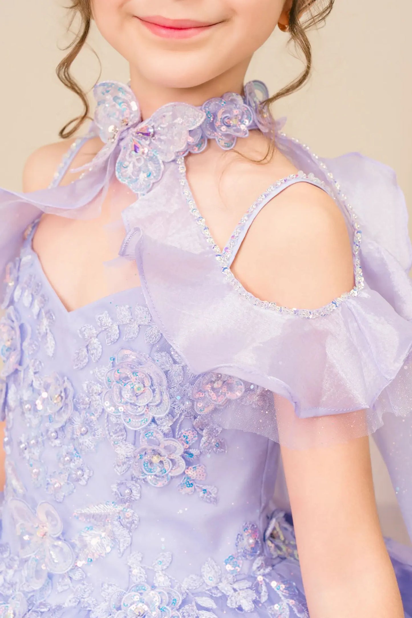 Side close up view of a kid wearing lilac mini quinceañera dress highlighting layered tulle skirt, ruffled off-shoulder sleeves, and sparkling embellished bodice.