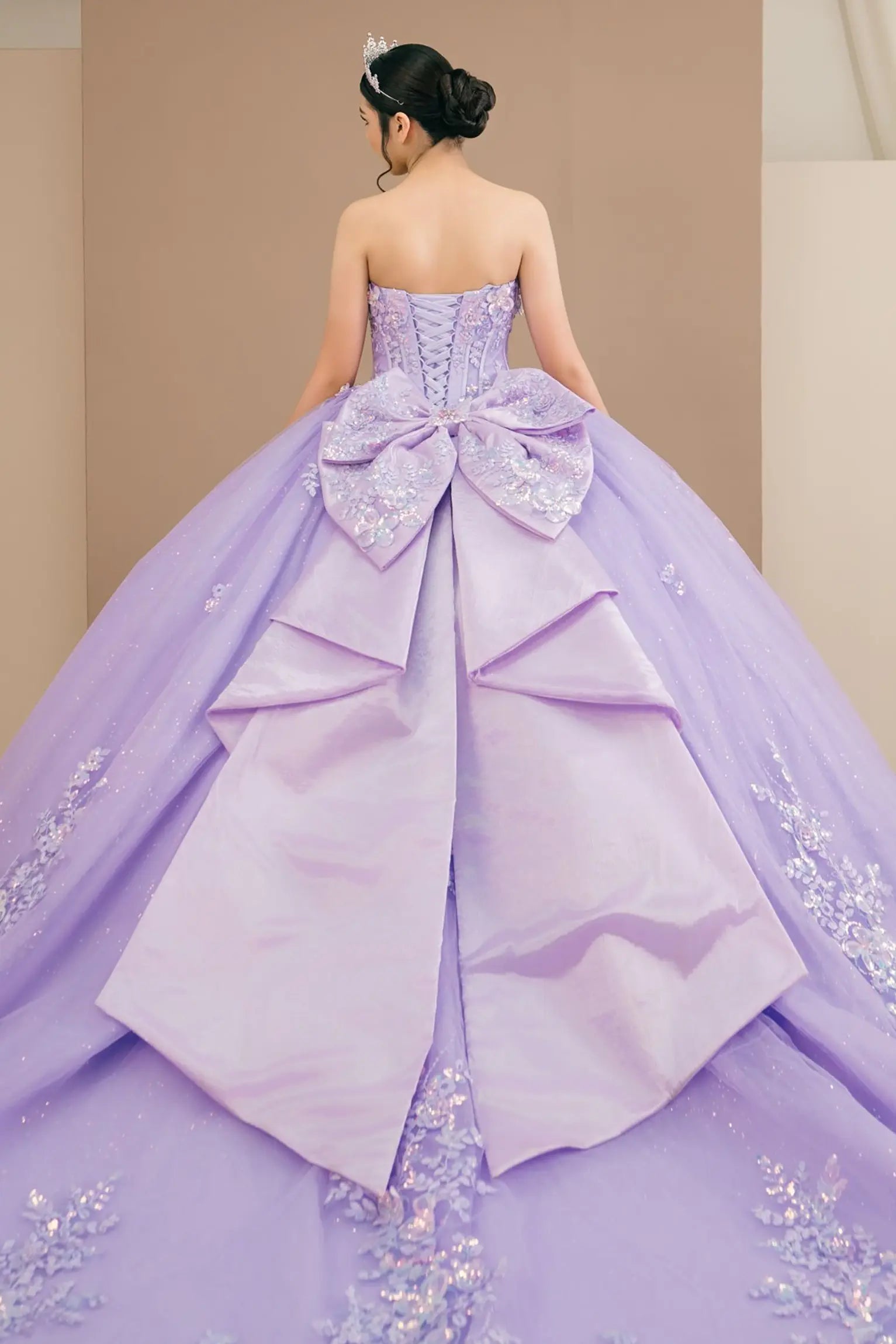 Back view of a woman wearing lilac quinceañera dress with voluminous tulle skirt, lace-up corset closure, and floral appliques trailing across the bodice.