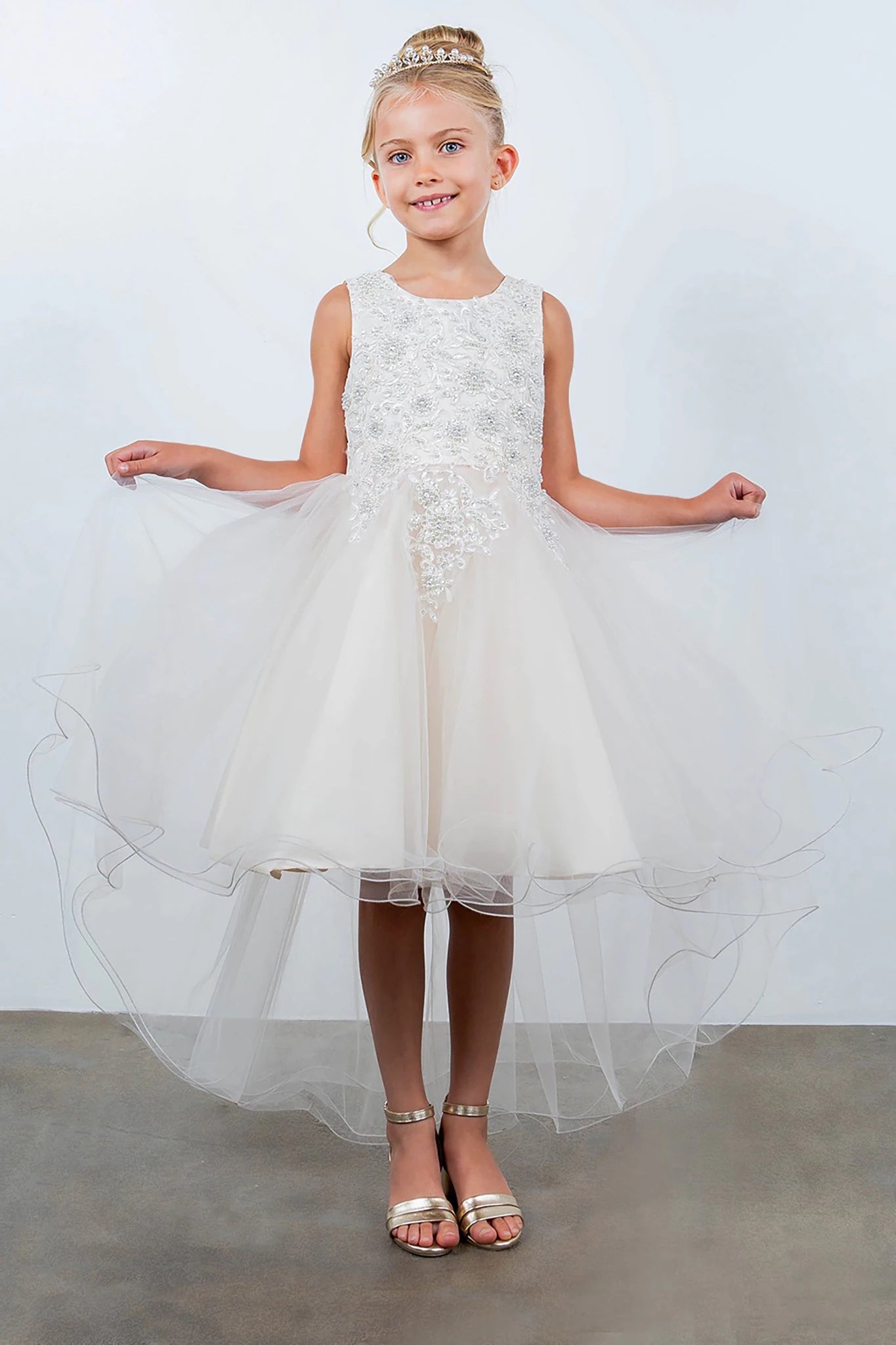 Front view of champagne sleeveless high-low tulle flower girl dress with lace bodice, pearl trim, rhinestone embellishments, and layered skirt.