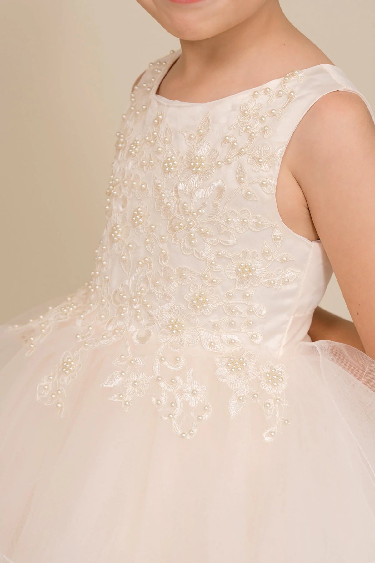 Close-up front-facing view of champagne sleeveless high-low tulle flower girl dress with lace bodice, pearl trim neckline, and rhinestone detailing.