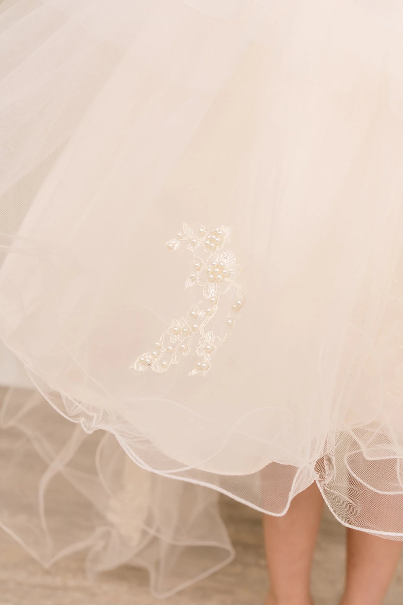Close-up of champagne sleeveless high-low tulle flower girl dress fabric with lace embroidery, pearl trim accents, rhinestone sparkle, and shimmering tulle layers.