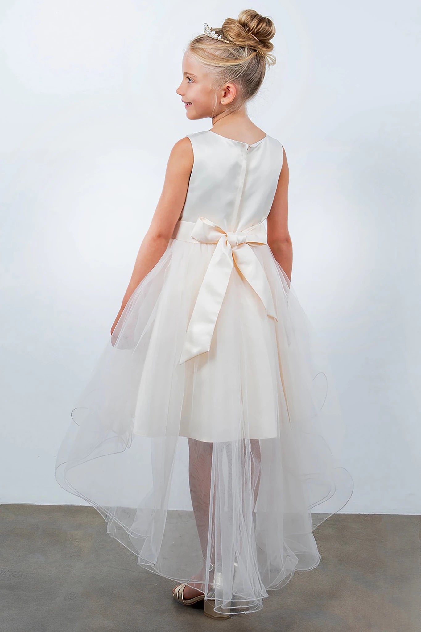 Back view of champagne sleeveless high-low tulle flower girl dress with lace-up closure, pearl trim neckline, rhinestone details, and cascading skirt.
