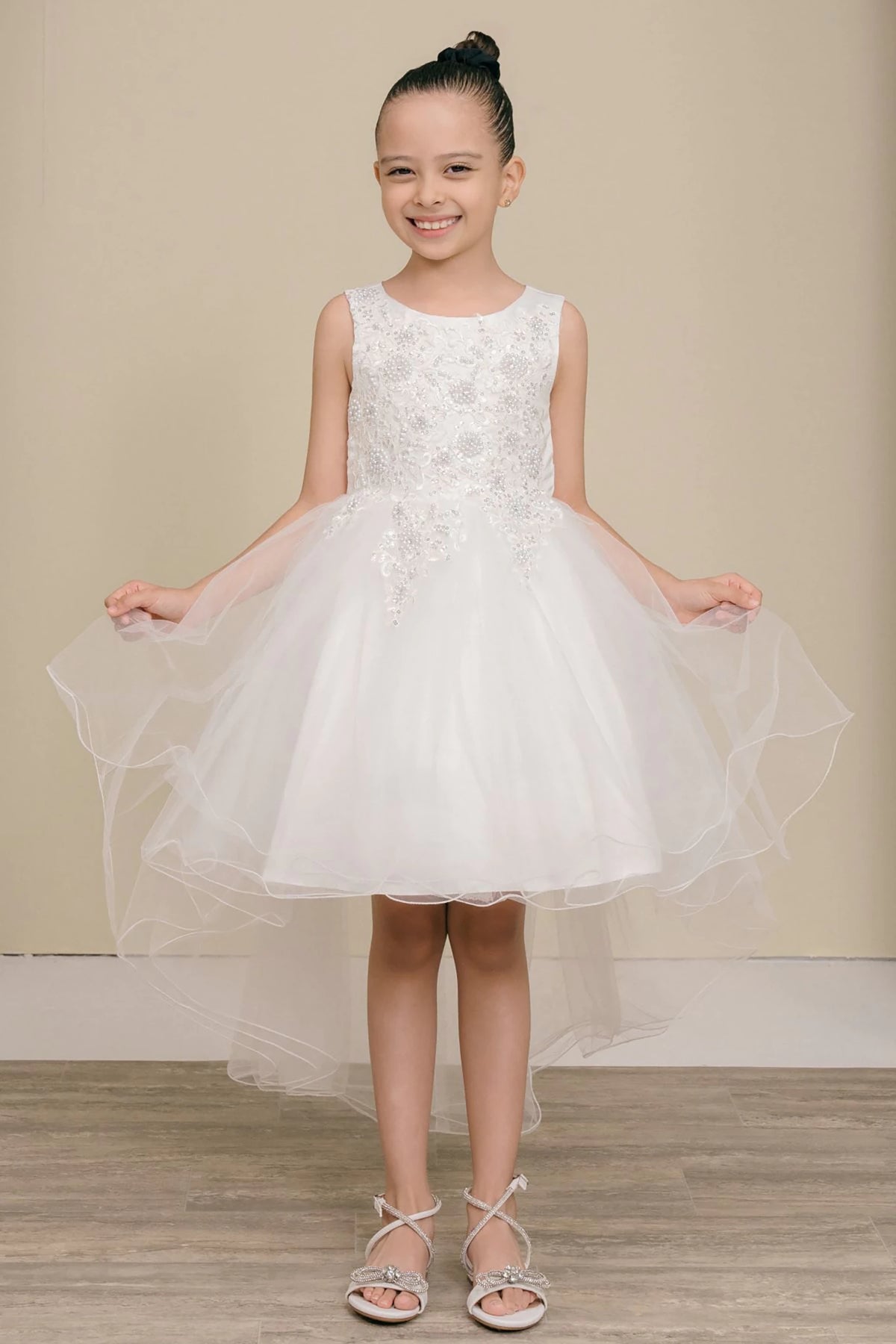 Front view of white sleeveless high-low tulle flower girl dress with lace bodice, pearl trim, rhinestone embellishments, and layered skirt.