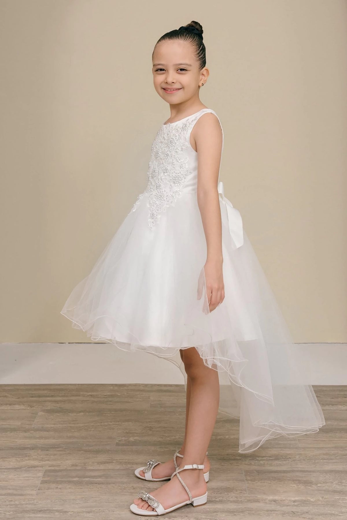 Side view of white sleeveless high-low tulle flower girl dress with lace detailing, pearl accents, rhinestone sparkle, and flowing layered skirt.