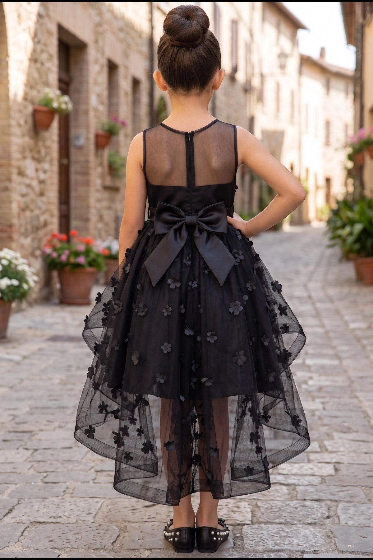 Displayed in a back view, the Black flower girl dress has sheer back, large bow and floral applique high-low train.