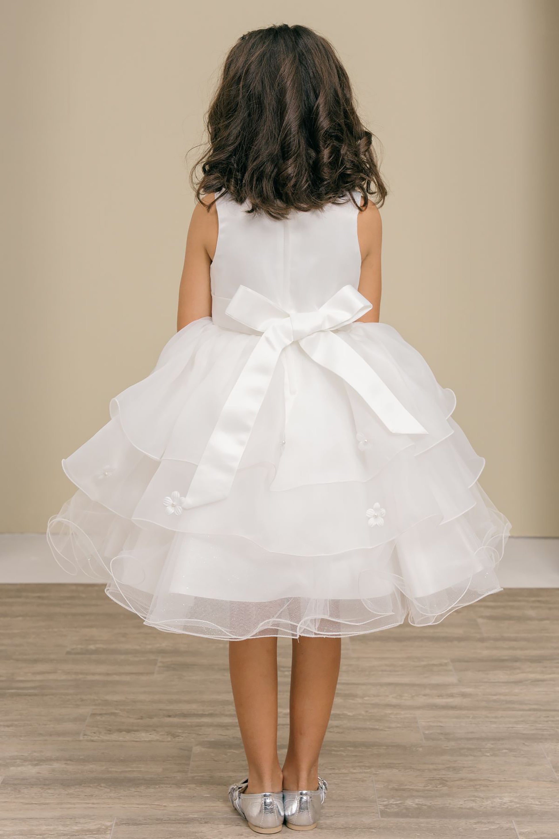 Young girl wearing a white dress with a large bow at the back against a beige wall.