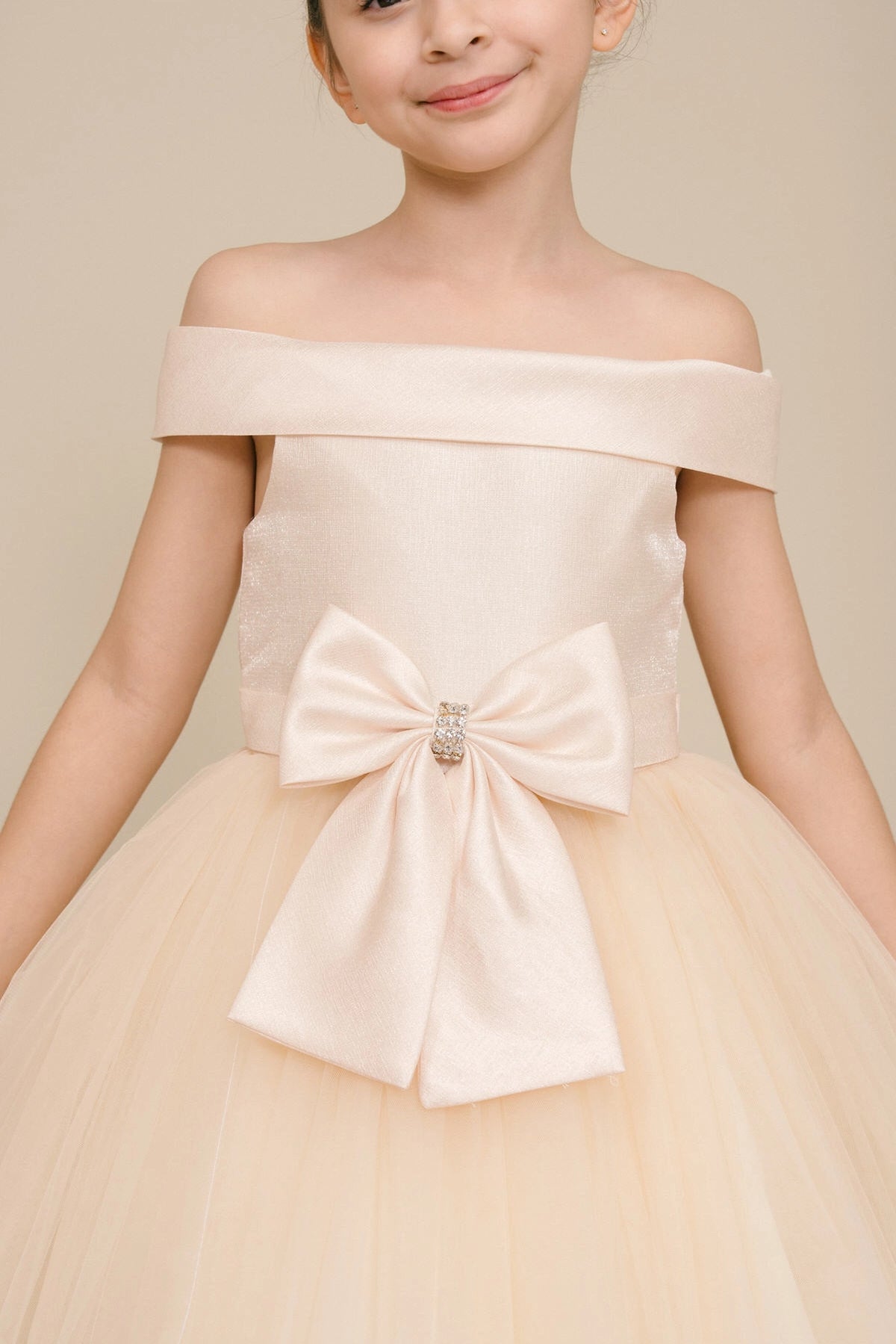 Visible on this Champagne flower girl dress: jeweled front bow beneath the off-shoulder neckline.