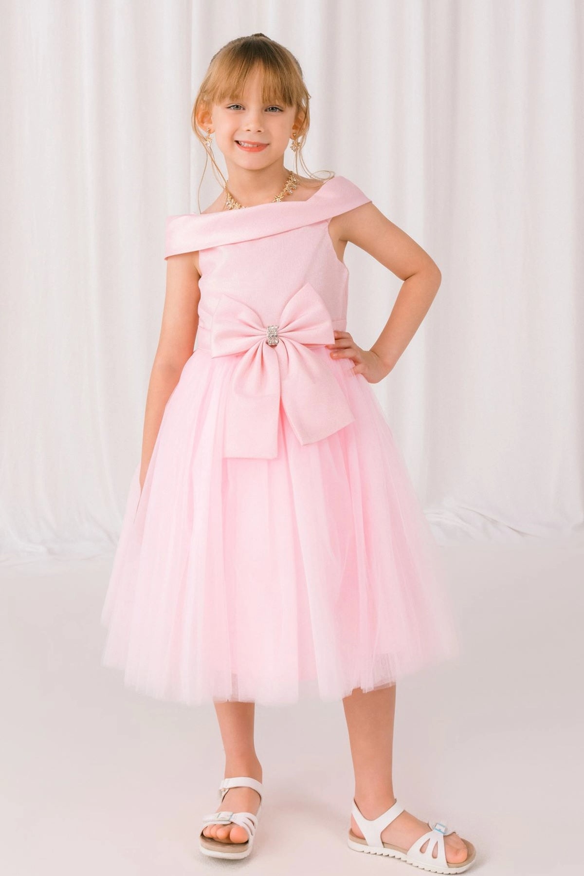 Pink flower girl dress features off-shoulder neckline and front jeweled bow.