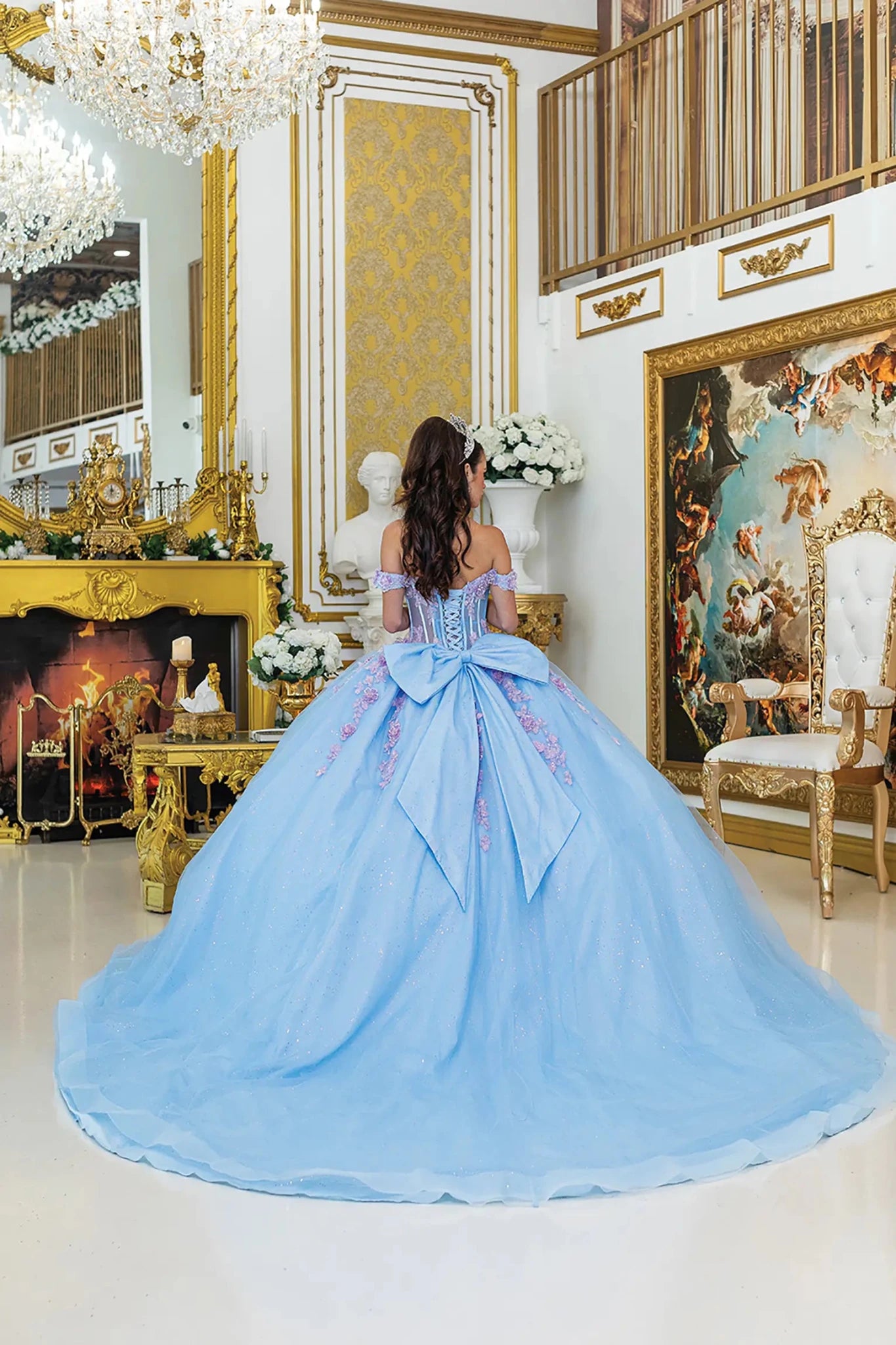 Woman standing back-facing in Bahama-lilac quinceañera ball gown with lace-up corset bodice, off-shoulder floral details, 3D embellishments, and flowing glitter tulle skirt with train