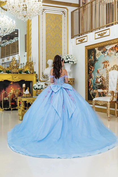 Woman standing back-facing in Bahama-lilac quinceañera ball gown with lace-up corset bodice, off-shoulder floral details, 3D embellishments, and flowing glitter tulle skirt with train