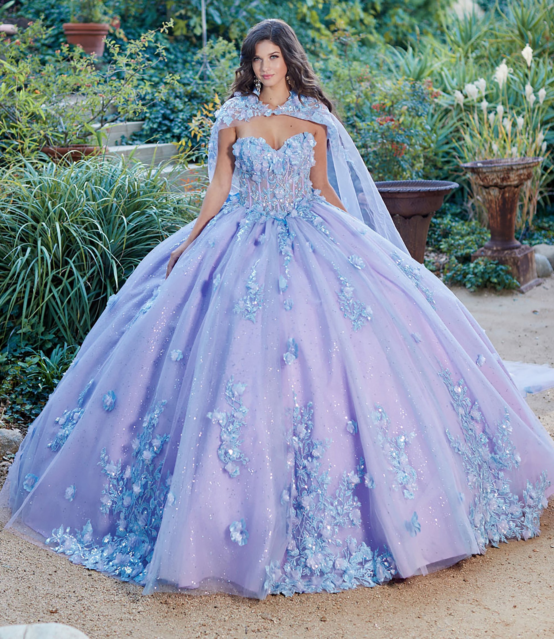 Quinceanera | Detachable Puff Sleeves and Cape Sparkly Ball Gown | Lilac Two Tone 3D Floral Detail Quince Dress - front view of woman wearing dress with one hand near her chin looking into the camera