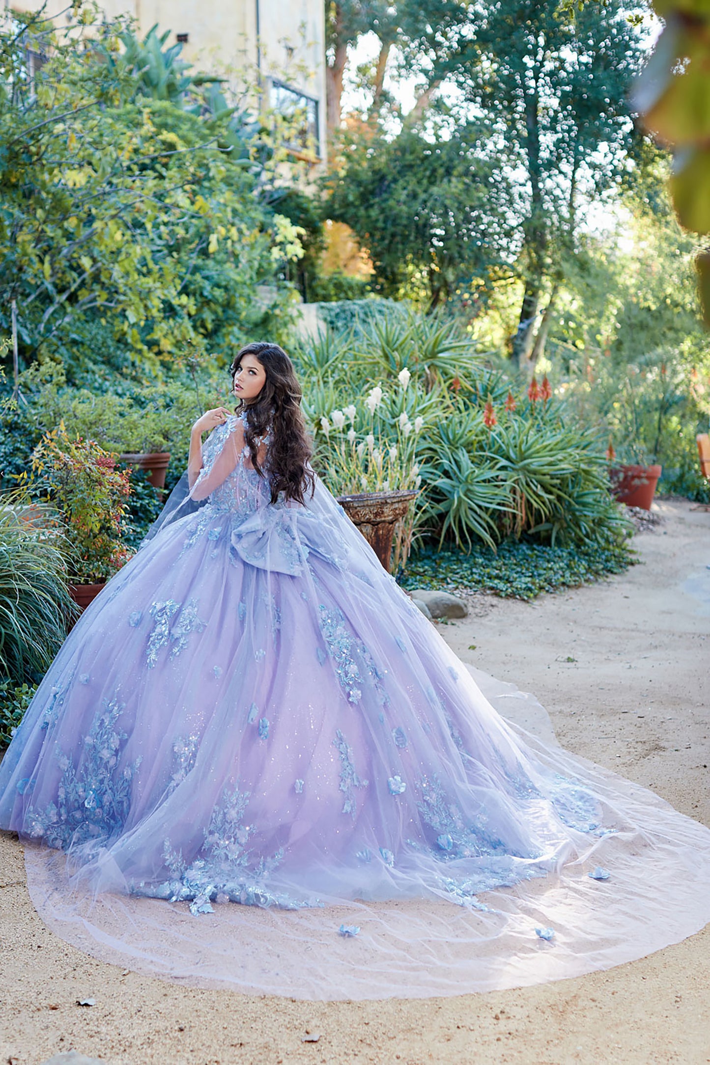 Quinceanera | Detachable Puff Sleeves and Cape Sparkly Ball Gown | Lilac Two Tone 3D Floral Detail Quince Dress - back view of woman wearing dress with her head turned back looking into the camera