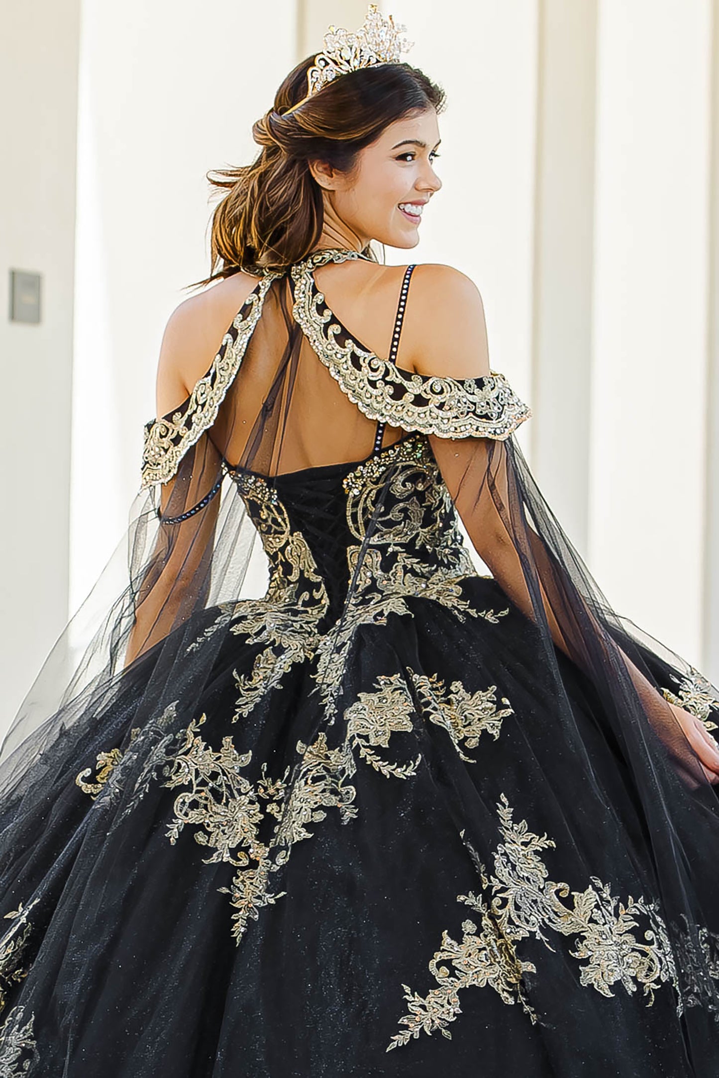 Quinceanera | Black with Gold Embroidered Lace with Stunning Cape Quince Ballgown dress - back view of woman wearing dress and crown smiling