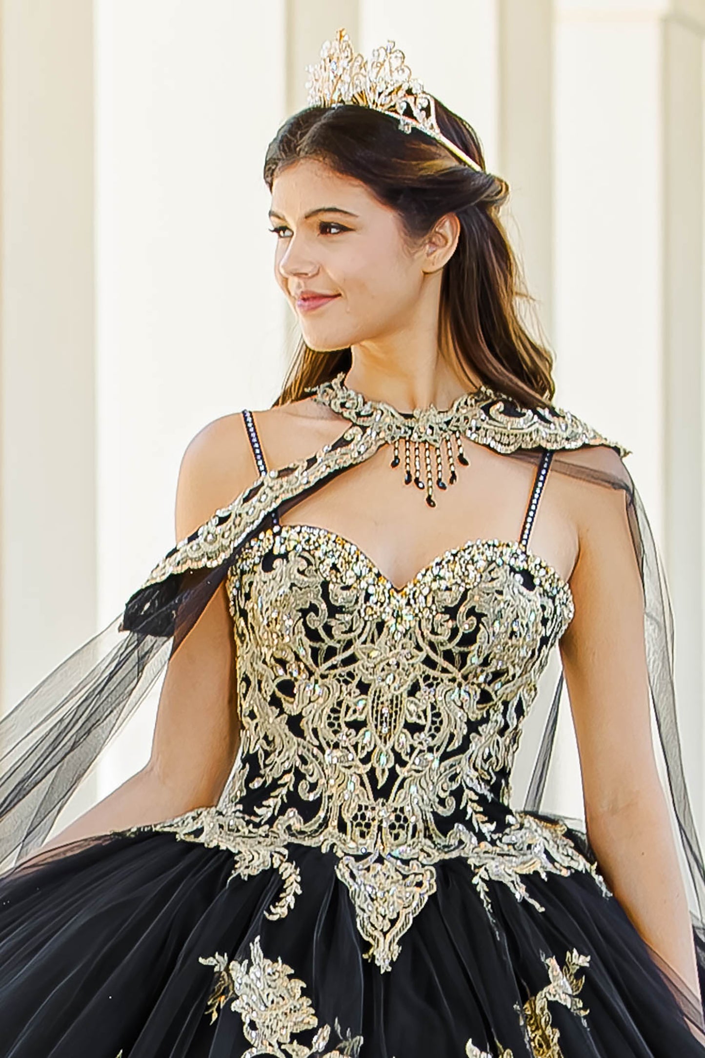 Quinceanera | Black with Gold Embroidered Lace with Stunning Cape Quince Ballgown dress - zoomed in front view of woman wearing dress and crown smiling look off into the distance