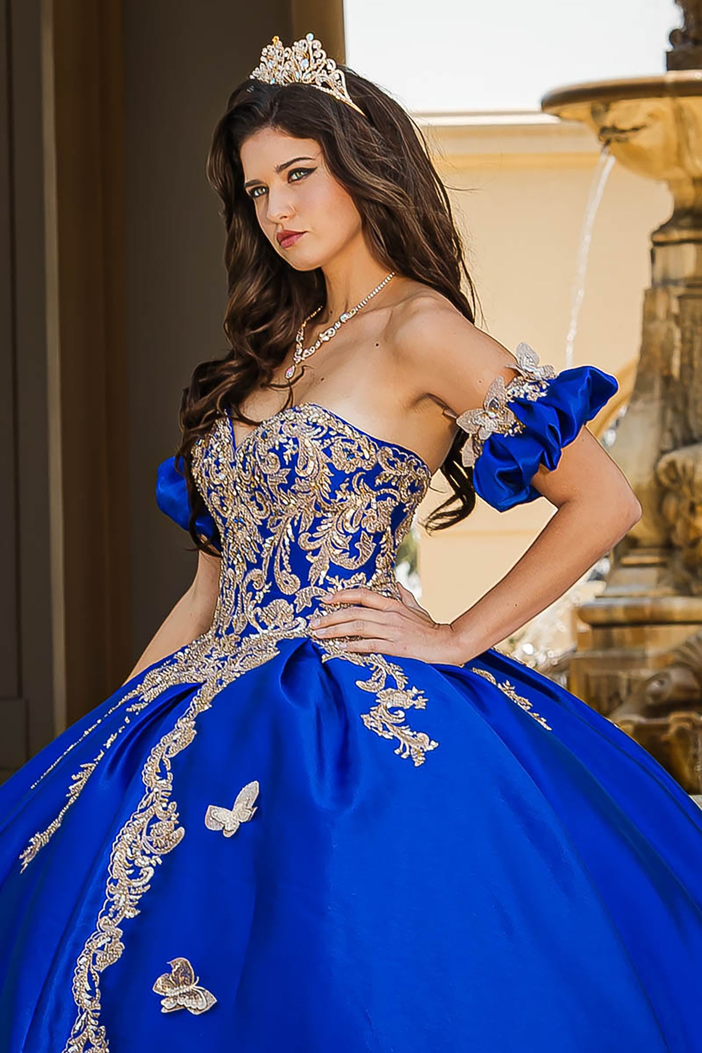 Woman in a blue and gold quinceañera dress with a tiara, standing in front of a fountain.