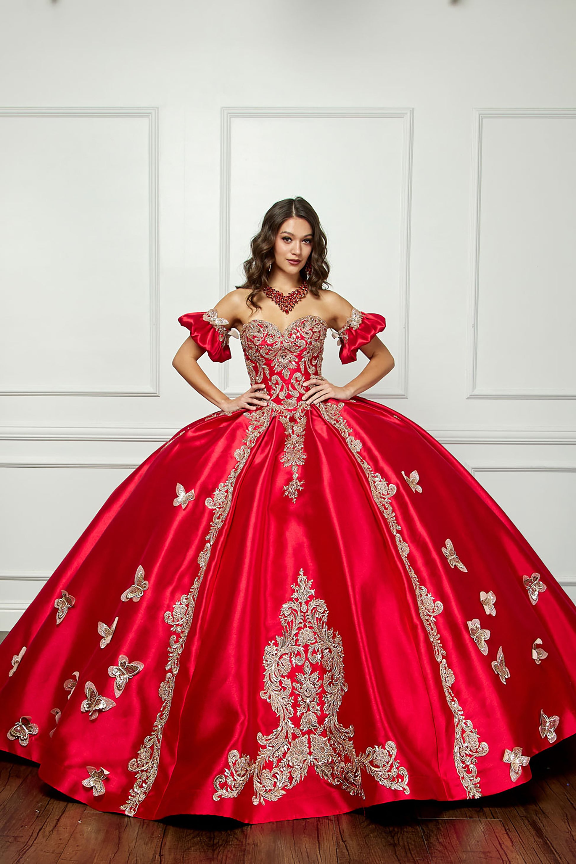 a girl wearing a red satin quinceanera ball gown with gold embroidery on a white wall background. the dress is fully satin.