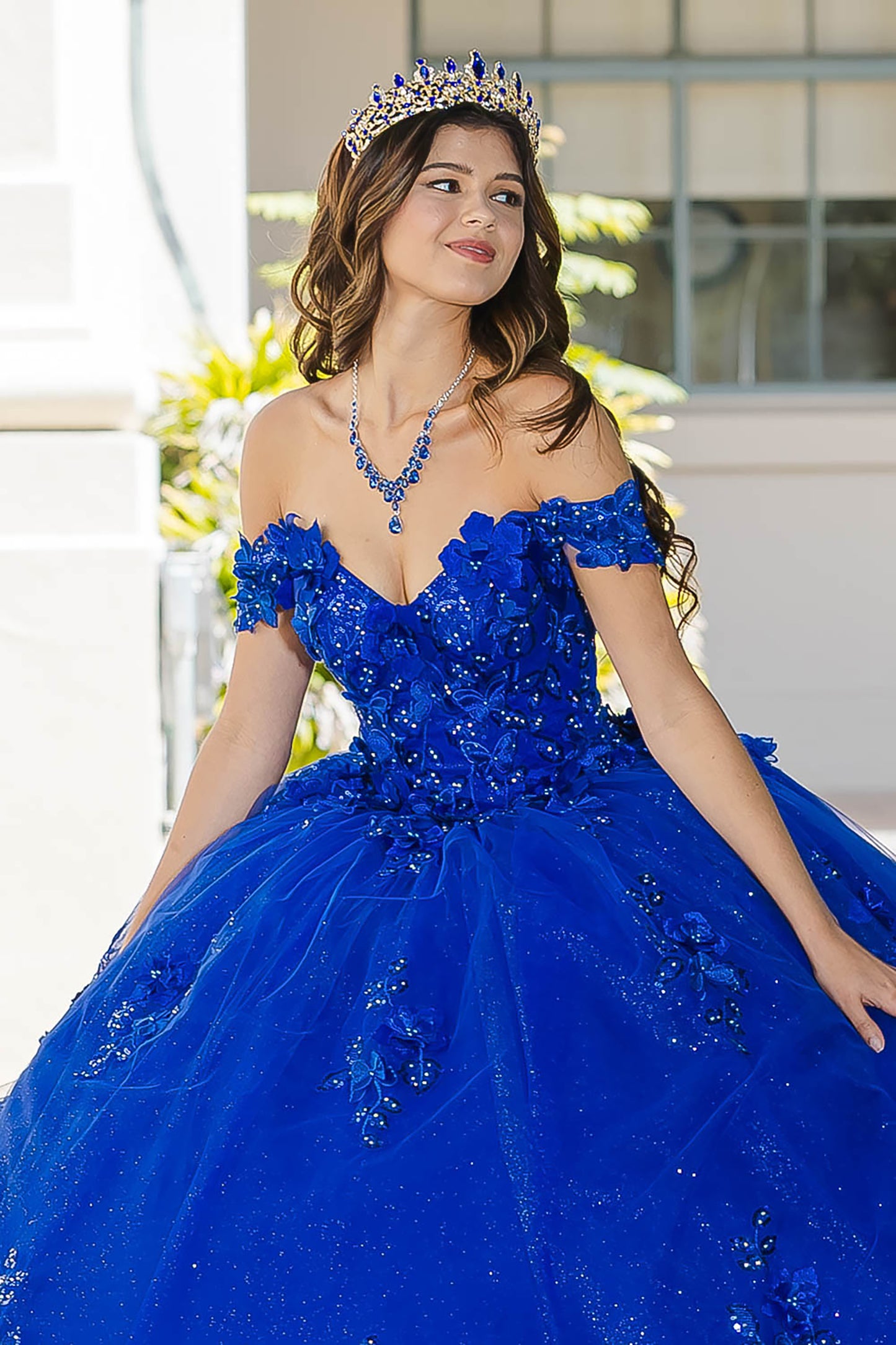 Blue Long Train Off-Shoulder Sequin detail 3D Flower and Butterflies bodice with Sparkle Tulle Quince Dress - front view of woman wearing dress and crown with hands at her side