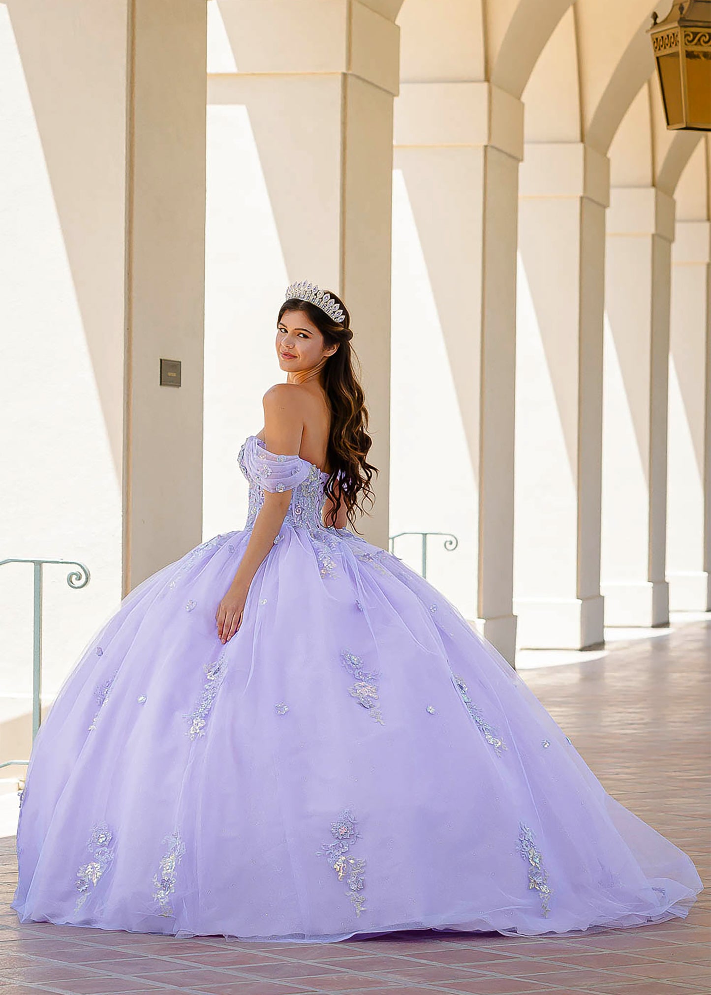 Quinceanera | Lilac Two-Tone See-Through Shimmering Sequins and 3D flowers Bodice Ballgown - back view of woman wearing dress and crown looking over her shoulder into the camera