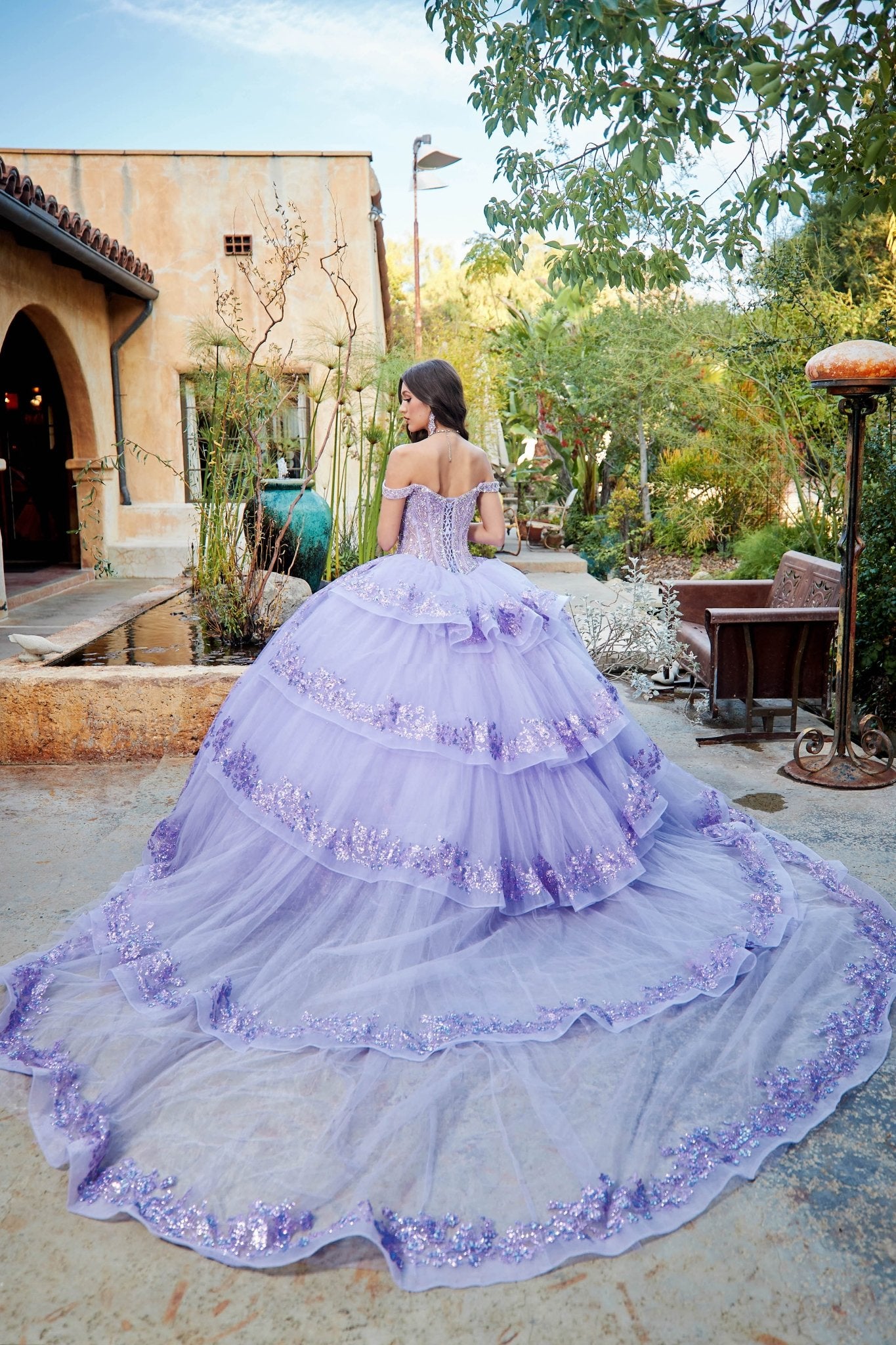 Woman in a lilac quince lace up back ball gown with an extra long ruffle train standing outdoors with greenery and a building in the background