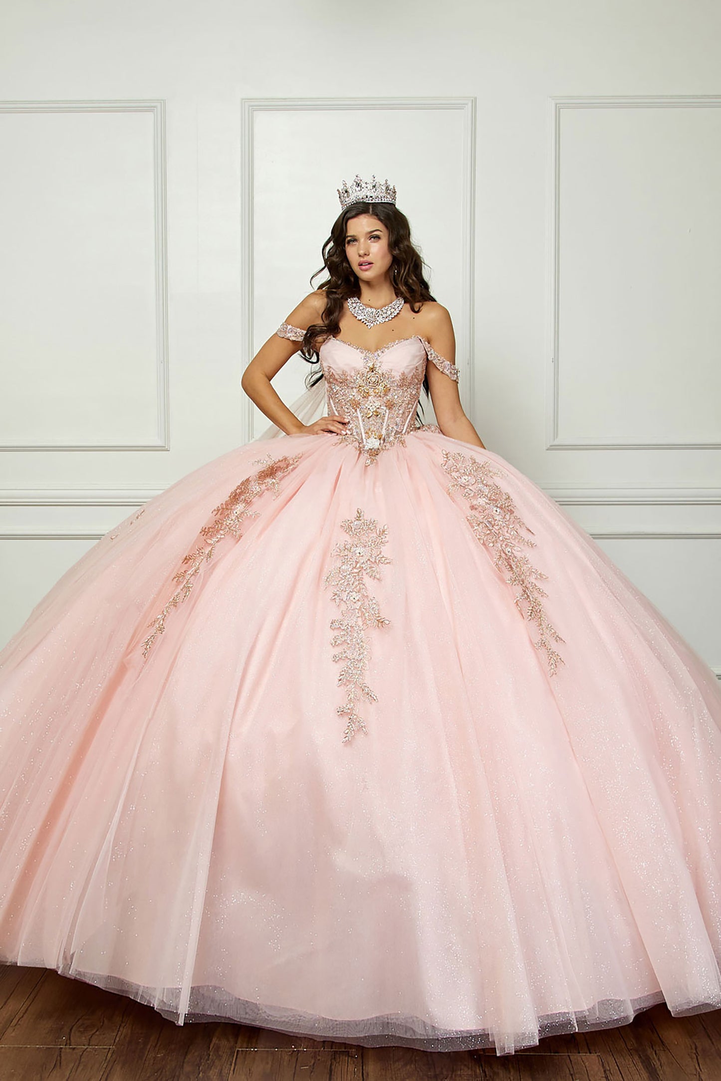 Quinceanera | Blush Off-Shoulder Ballgown with Beading, Rhinestones, 3D Flowers, Golden Embroidery, and Detachable Glitter Cape - front view of woman wearing dress with cronw on head and hand on hip