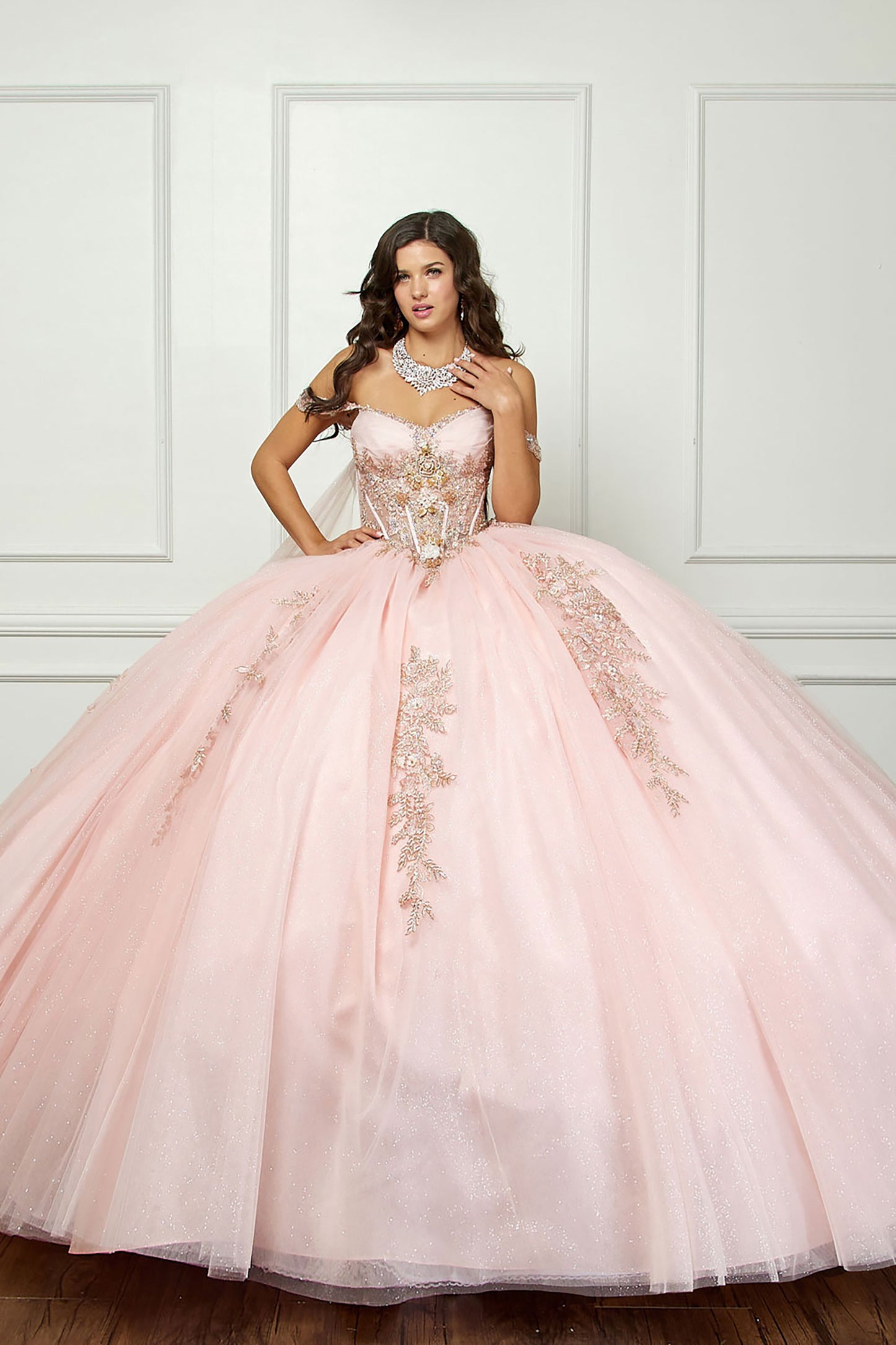 Quinceanera | Blush Off-Shoulder Ballgown with Beading, Rhinestones, 3D Flowers, Golden Embroidery, and Detachable Glitter Cape - front view of woman wearing dress with one hand on necklace and other hand on hip