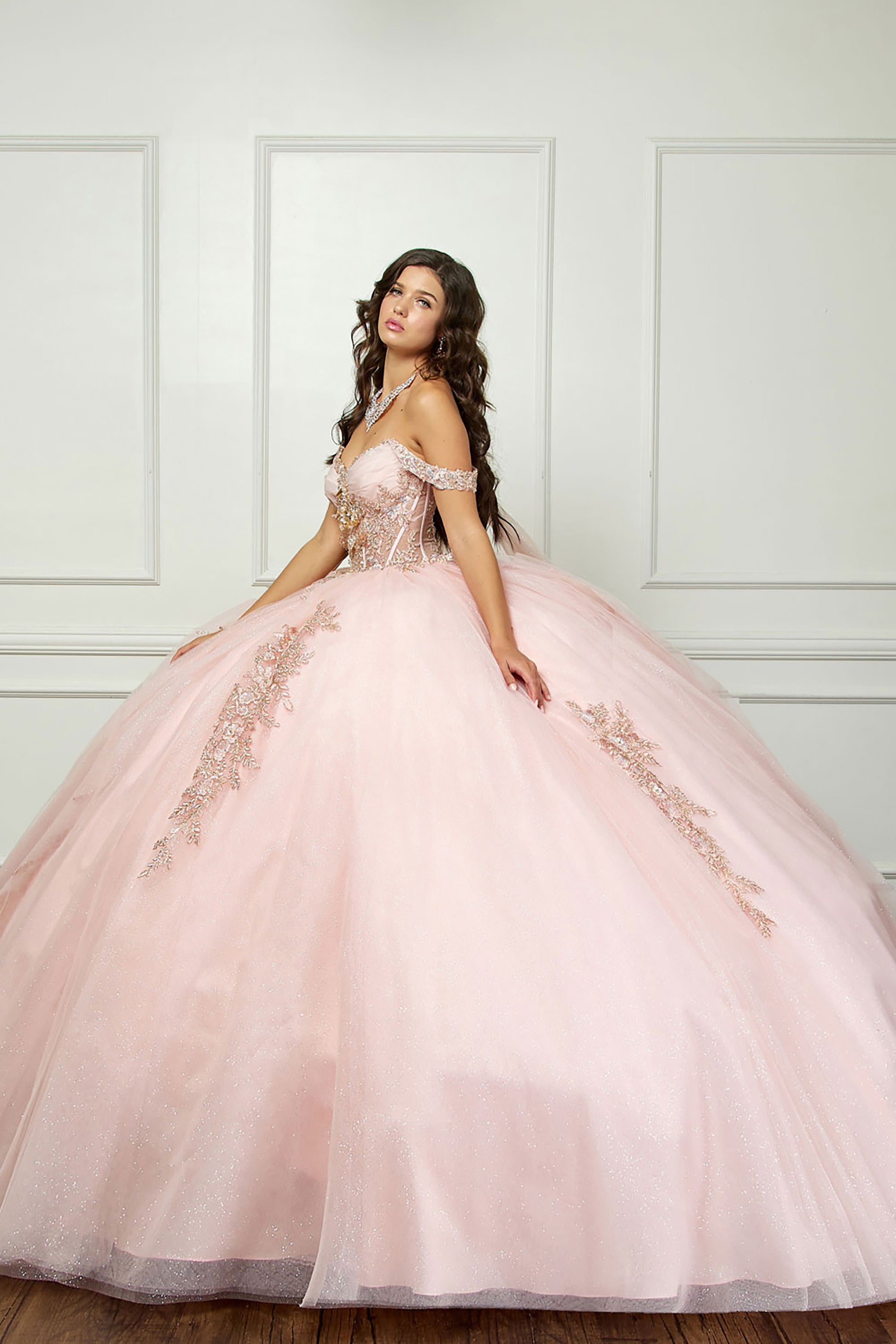 Quinceanera | Blush Off-Shoulder Ballgown with Beading, Rhinestones, 3D Flowers, Golden Embroidery, and Detachable Glitter Cape - side view of woman wearing dress with hand at side of skirt