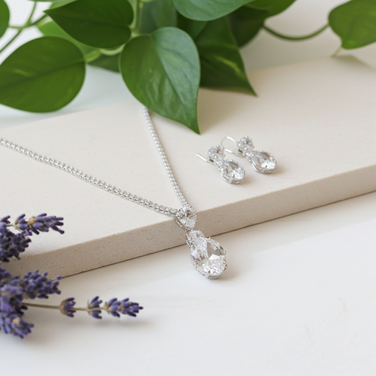 Silver rhinestone jewelry set with necklace and earrings on a white surface with green leaves and lavender flowers.