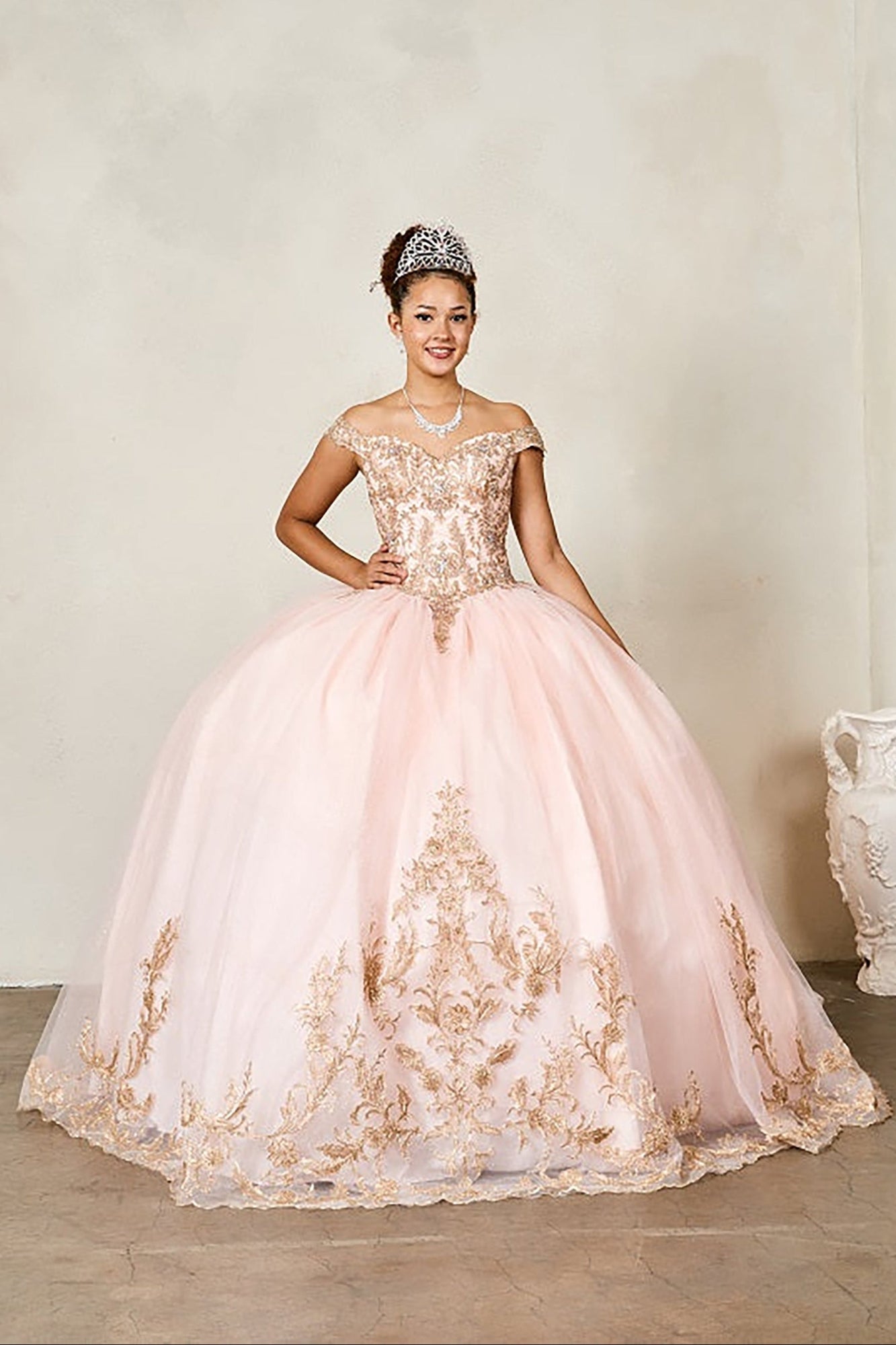 Woman in a pink and gold embroidered gown with a tiara, standing against a plain background.