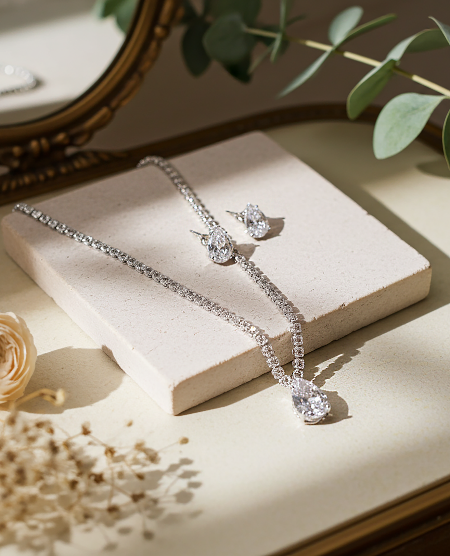 Silver rhinestone jewelry set with necklace and earrings on a light stone flatform, next to pretty dried wedding ivory flower, in front of pale green plant, the objects stand out as a silver jewelry

