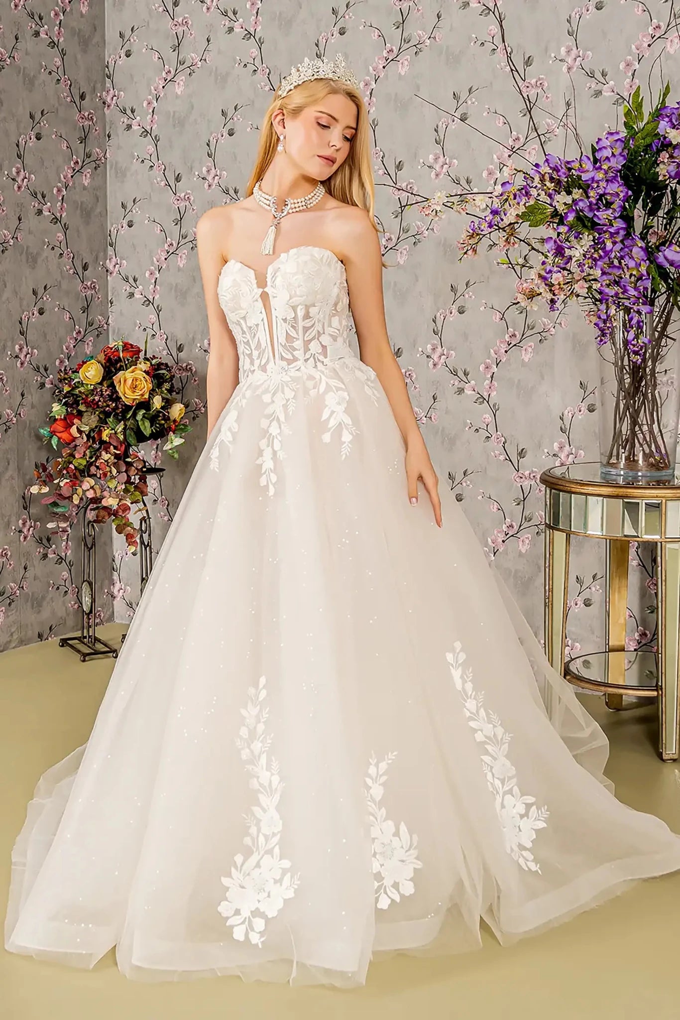 Front full-length view of an ivory ball gown featuring an illusion sweetheart neckline, a strapless sheer bodice, and a voluminous skirt layered with tulle and shimmering accents.