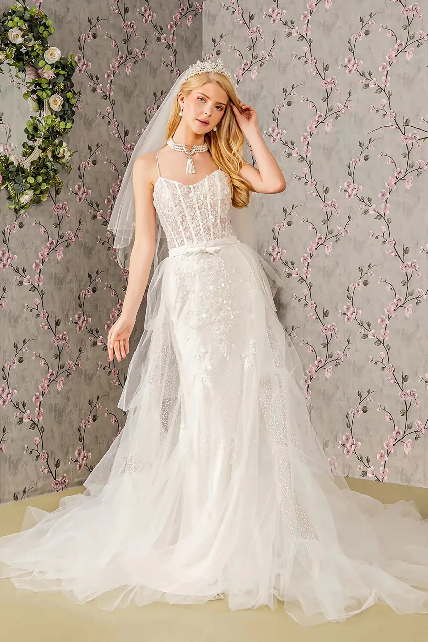 Front full-length view of an ivory mermaid wedding dress featuring a scoop neckline, spaghetti straps, and a long mesh cape flowing over an embroidered sequin and glitter body.