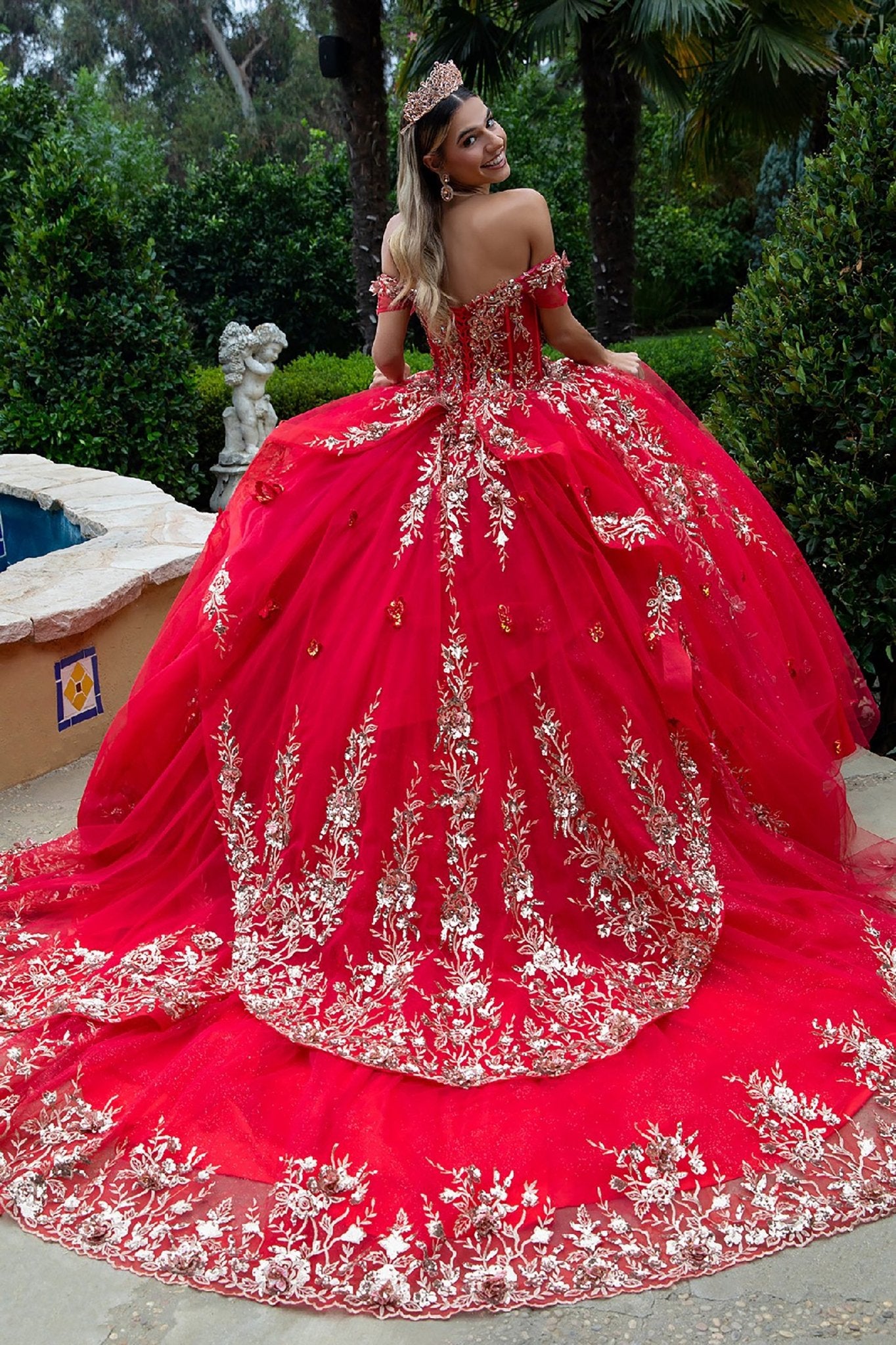Back view of ornate off-shoulder red gown with gold detailing and flowing train near fountain garden