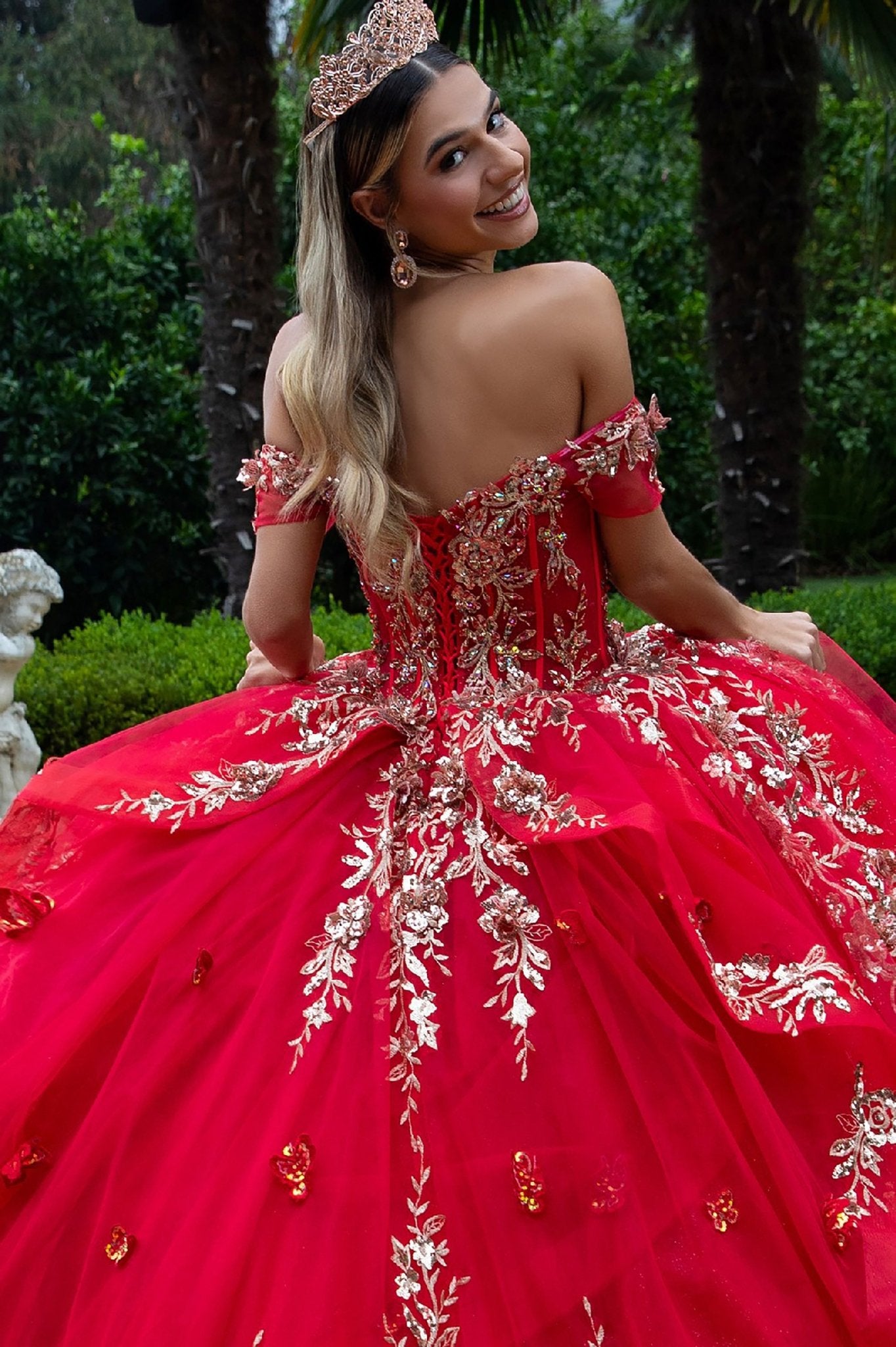 Back view of quinceañera red gown with gold embellishments and long train worn outdoors in tropical garden