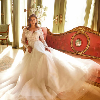 Front seated full-length view of an off-white A-line wedding dress featuring an illusion off-the-shoulder neckline, long embellished mesh sleeves, and a voluminous glitter tulle skirt.
