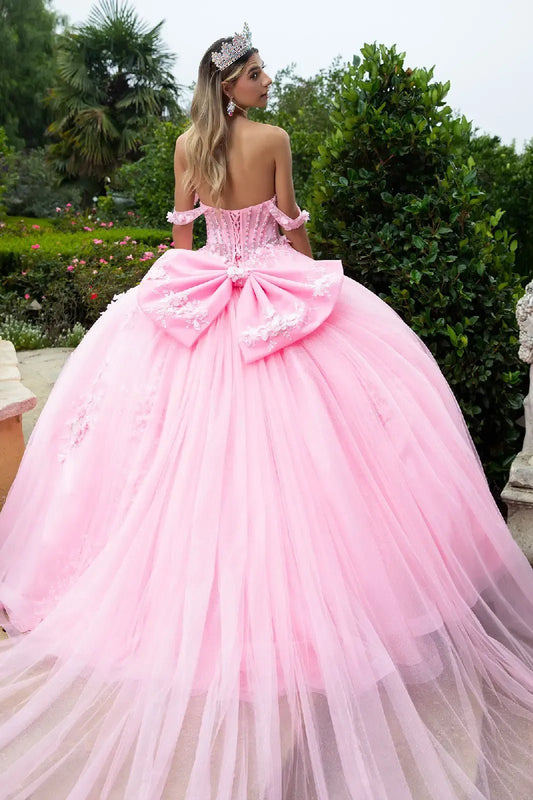 woman standing in a garden, wearing an elaborate floor‑length pink ball gown. The gown features a voluminous tulle skirt and a large decorative bow on the back, giving it a fairy‑tale elegance.