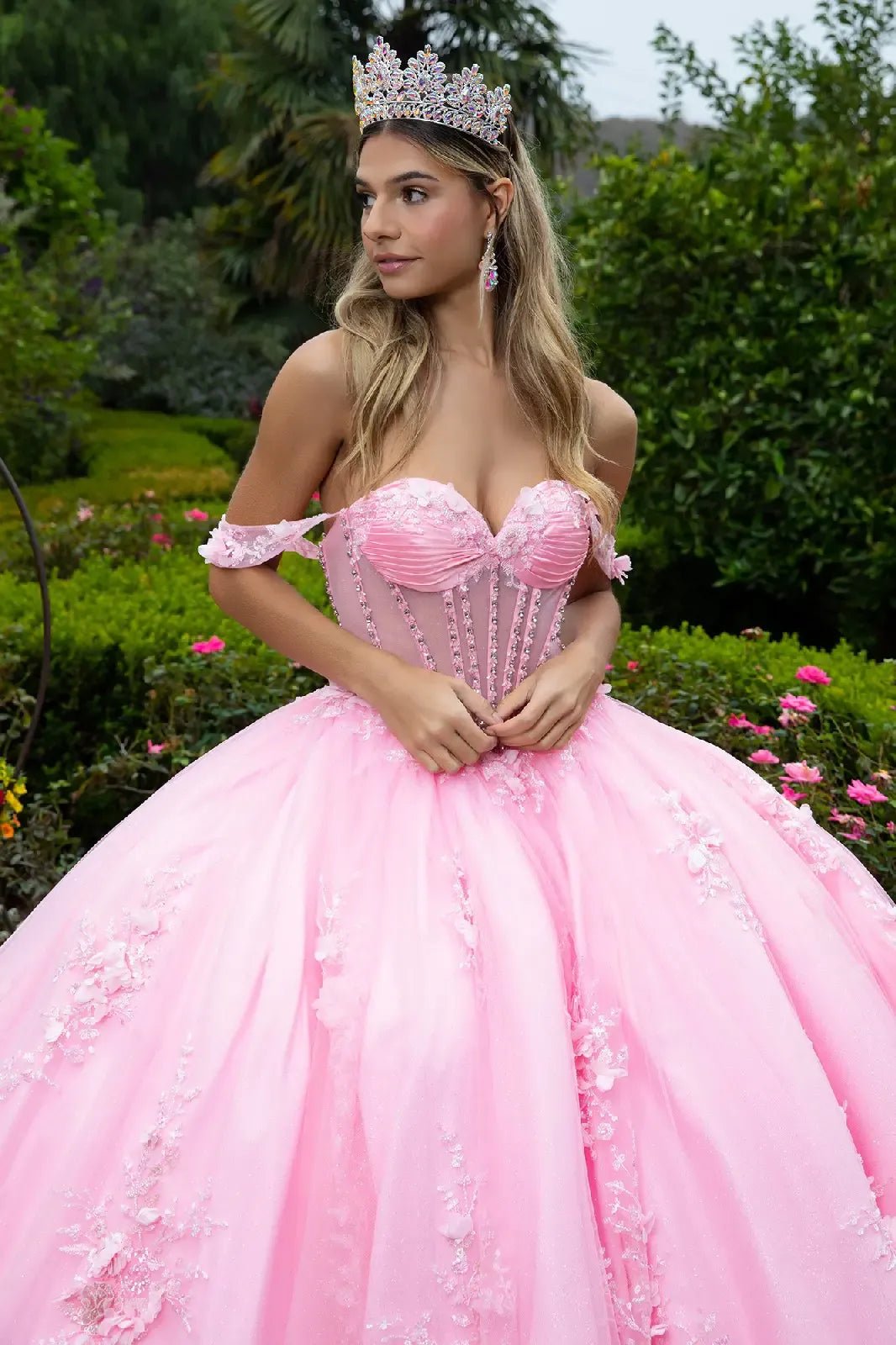 woman standing in a lush garden surrounded by greenery and blooming flowers. She is dressed in an elaborate strapless pink ball gown with a voluminous skirt.