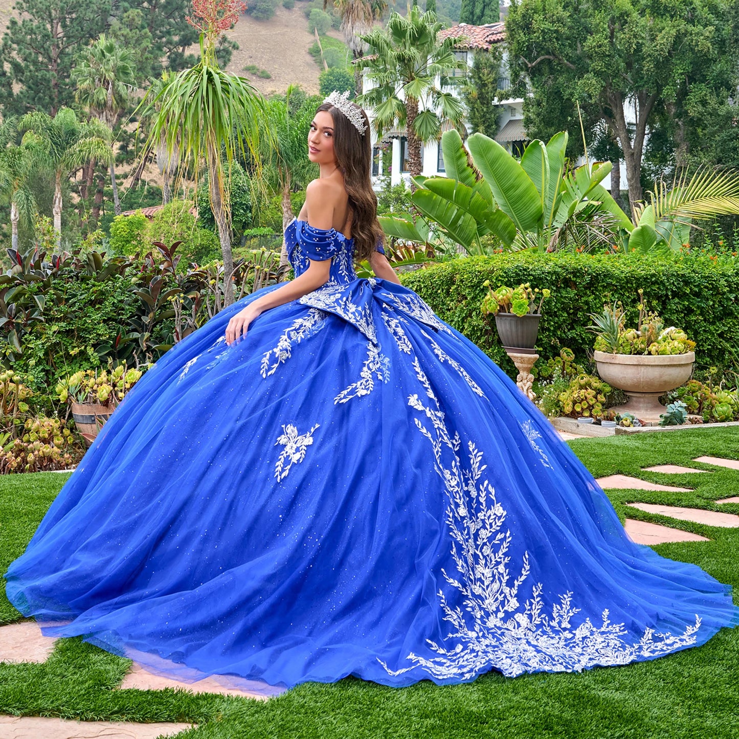 Woman in a blue ball gown with white embroidery standing in a garden.