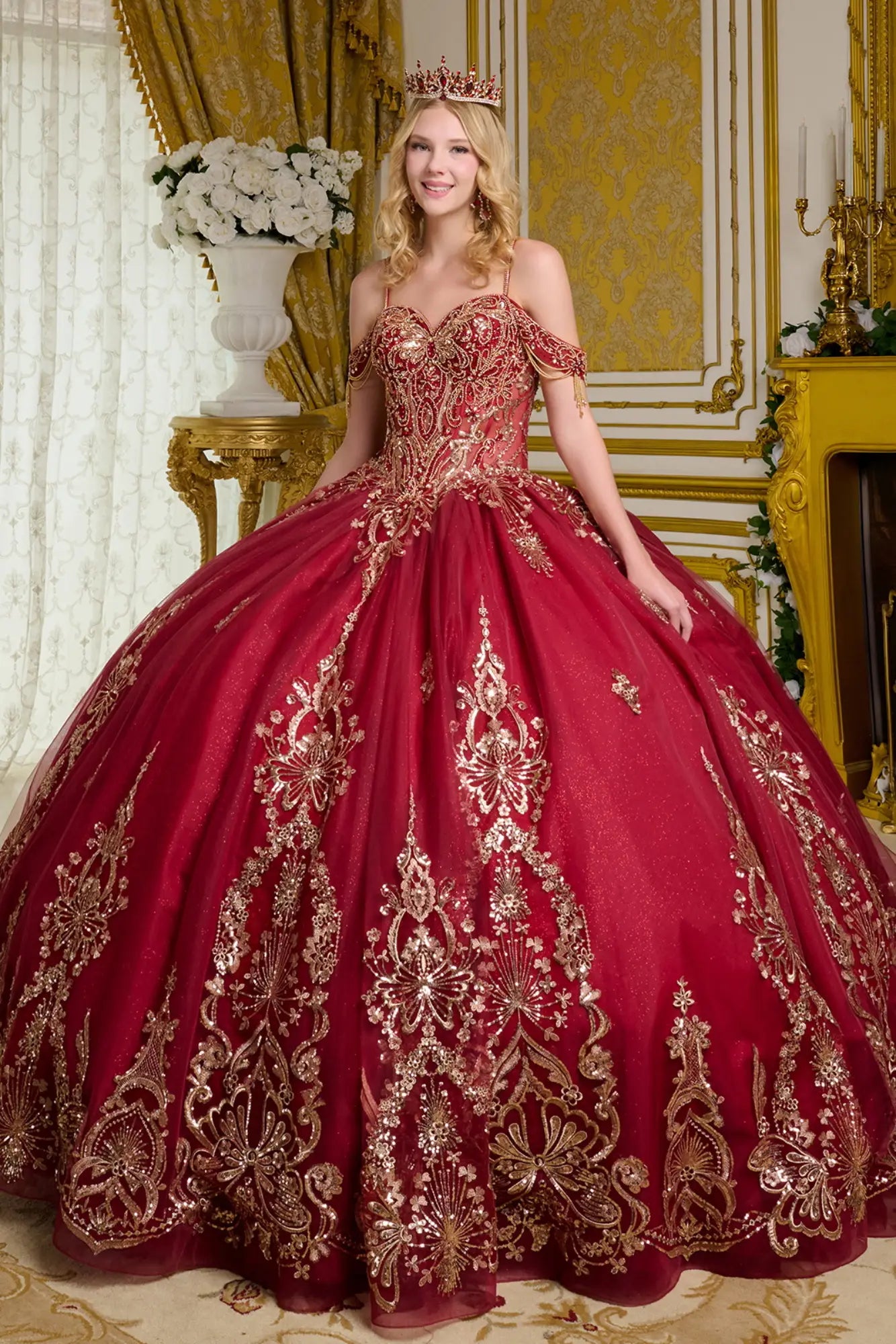 Front view of burgundy quinceañera ball gown with off-the-shoulder straps, heavily embroidered corset bodice, gold beadwork, and full glitter tulle skirt in an ornate palace interior