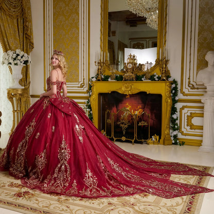 Wide back view of burgundy quinceañera ball gown with dramatic long train, gold embroidered patterns, detachable bow, and corset bodice in a luxury room setting
