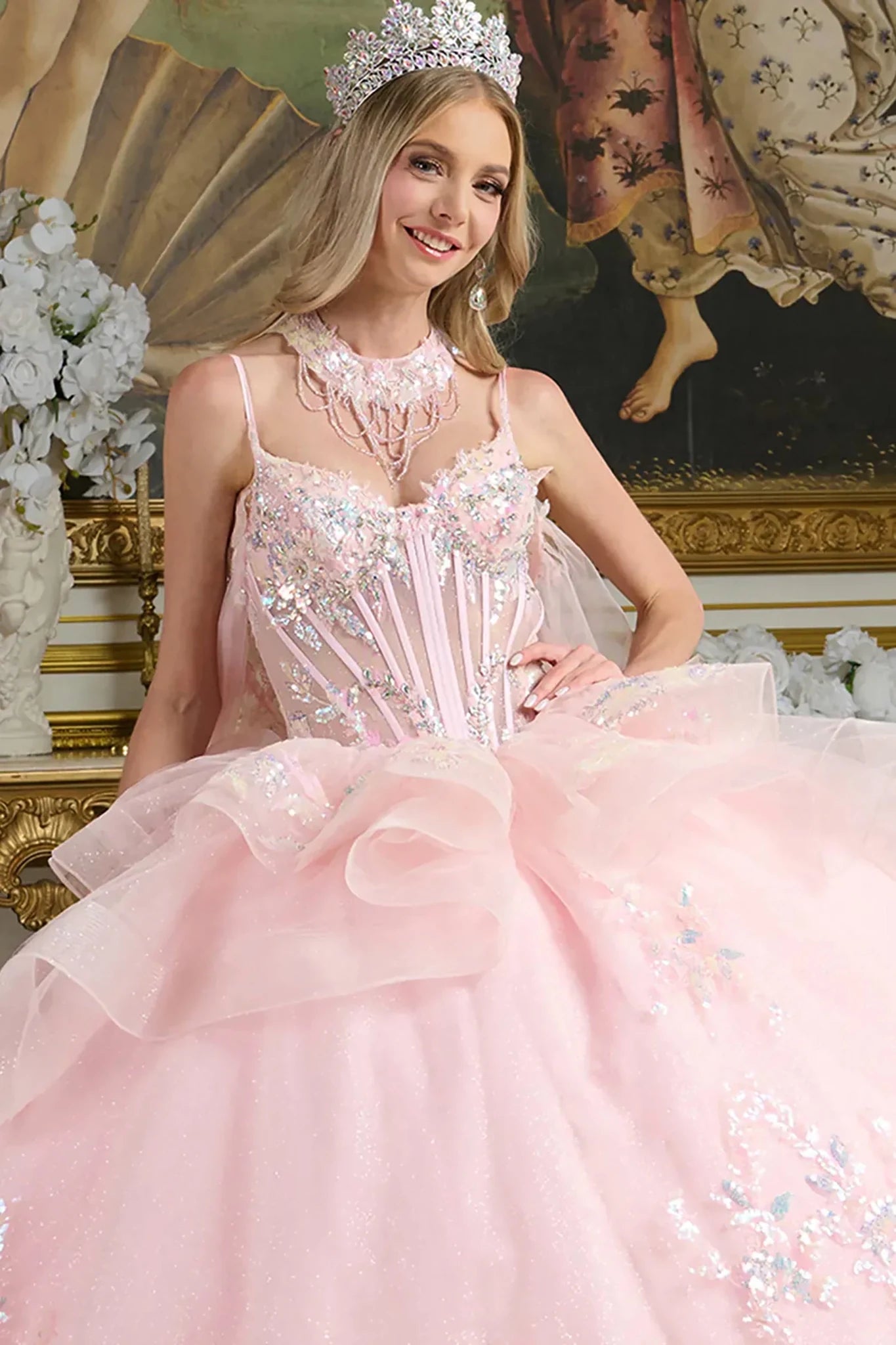 Woman in a pink gown with floral details and a tiara, sitting in an ornate room.